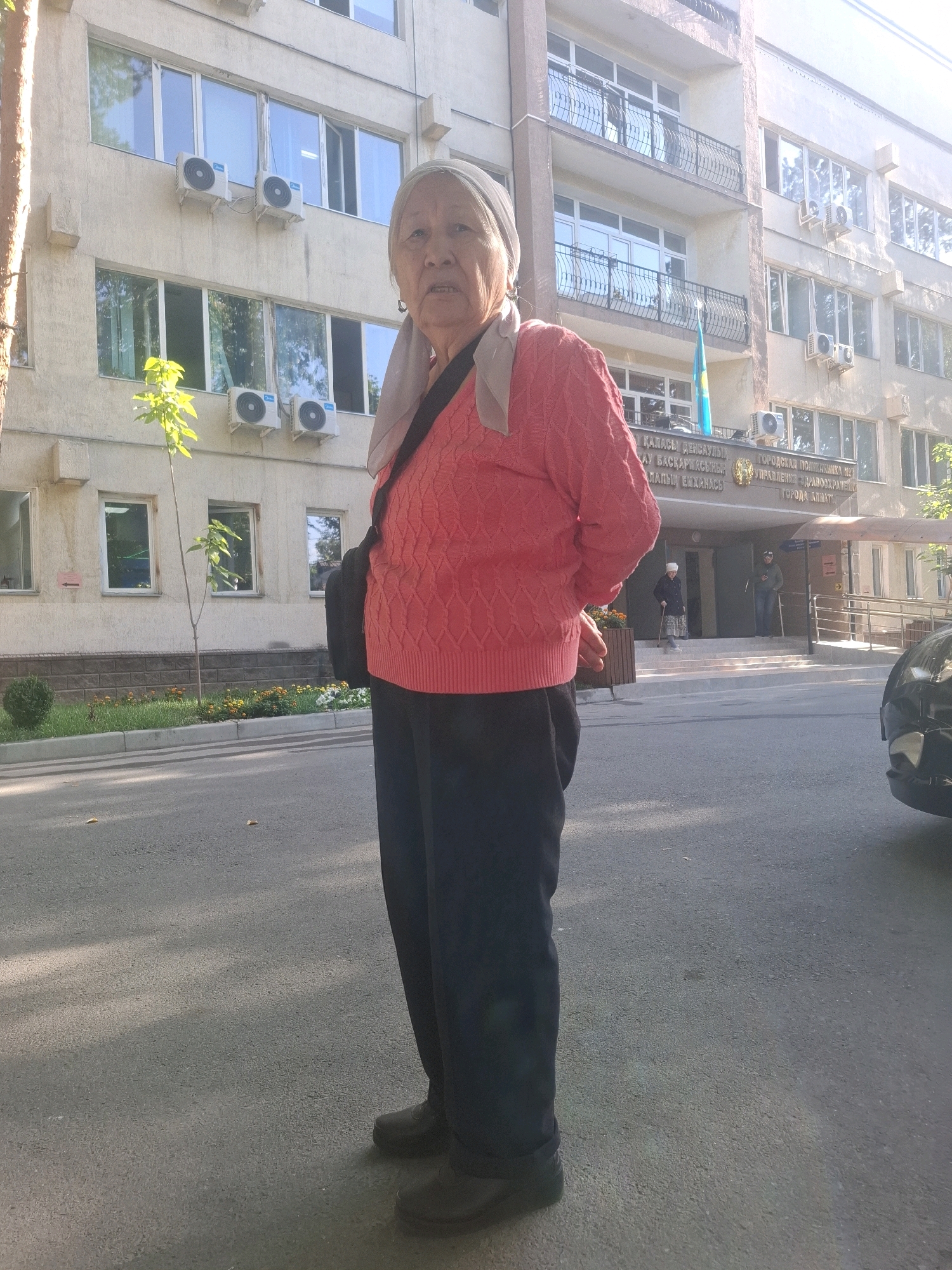 An elderly woman stands on an asphalt road, facing the camera. She wears a light-colored headscarf, a bright pink patterned sweater, and dark trousers. Her left hand is visible near her waist. Behind her, a light-colored building with multiple windows and air conditioning units is prominent. A small tree with green leaves stands to the left of the building entrance. The building has signs in Cyrillic script, and a flag of Kazakhstan is visible on a flagpole on the building's facade. Two people are seen standing at the entrance of the building, one of whom is using a cane. A black car is partially visible on the right side of the image. The scene appears to be a street in Almaty, Kazakhstan.
