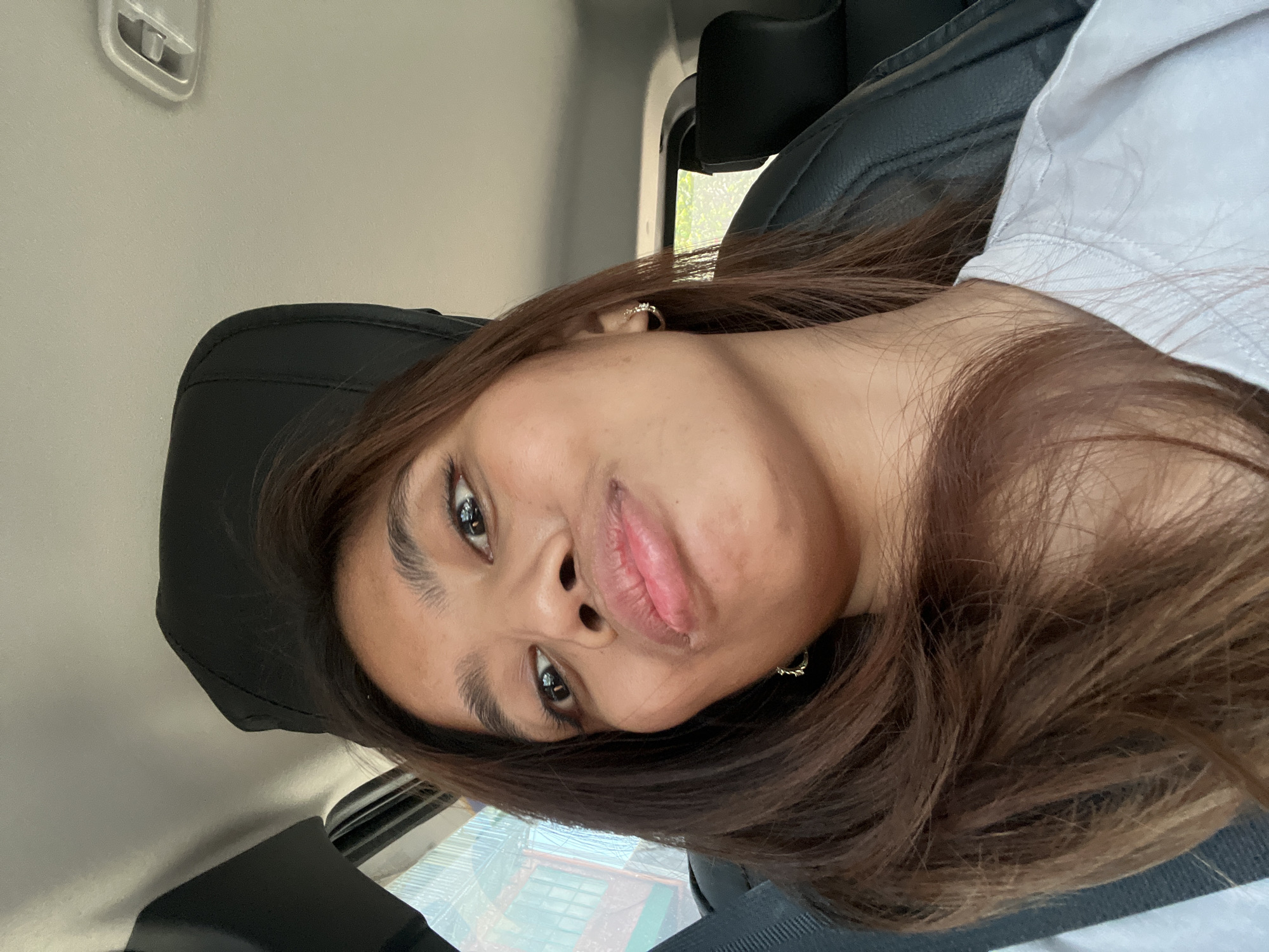 A young adult individual, likely female, with medium-toned skin and long, dark brown hair with lighter auburn streaks, is depicted in a low-angle selfie within a vehicle. Her head is tilted slightly down, and her eyes gaze upward and to the right. She exhibits a playful expression with pursed lips. She is wearing a white crew-neck t-shirt and small, gold-toned hoop earrings with white stone embellishments.
The interior of the vehicle includes visible black leather or synthetic seat upholstery, including headrests, and a dark grey seatbelt strap on the left side of the frame. The vehicle's ceiling is light-colored, featuring a rectangular overhead light fixture on the right. Through the left-side window, a blurred exterior scene reveals a building or structure with rectangular windows. The right-side window shows blurred green foliage. The scene is illuminated by natural daylight.
Based solely on the visual information provided, there are no discernible features or landmarks that permit identification of the specific location as Davao City, Philippines.