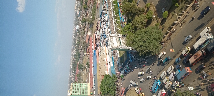 This high-angle image depicts a densely populated urban transportation area in Nairobi, Kenya, specifically the Machakos Country Bus Station vicinity. The foreground and mid-ground feature a multi-lane road network, including a roundabout, teeming with vehicles. A high concentration of buses and matatus (minibuses) occupies the roads and fills the expansive open-air bus station located centrally. Numerous pedestrians are observed moving on sidewalks and within the bus station area. To the far left, a prominent multi-story building with a light green facade stands against the urban skyline. An elevated metallic structure, likely a pedestrian walkway, bridges a section of the bus terminal. The background shows a broad expanse of the city under a clear, bright blue sky with sparse clouds. Scattered trees provide green elements within the built environment.