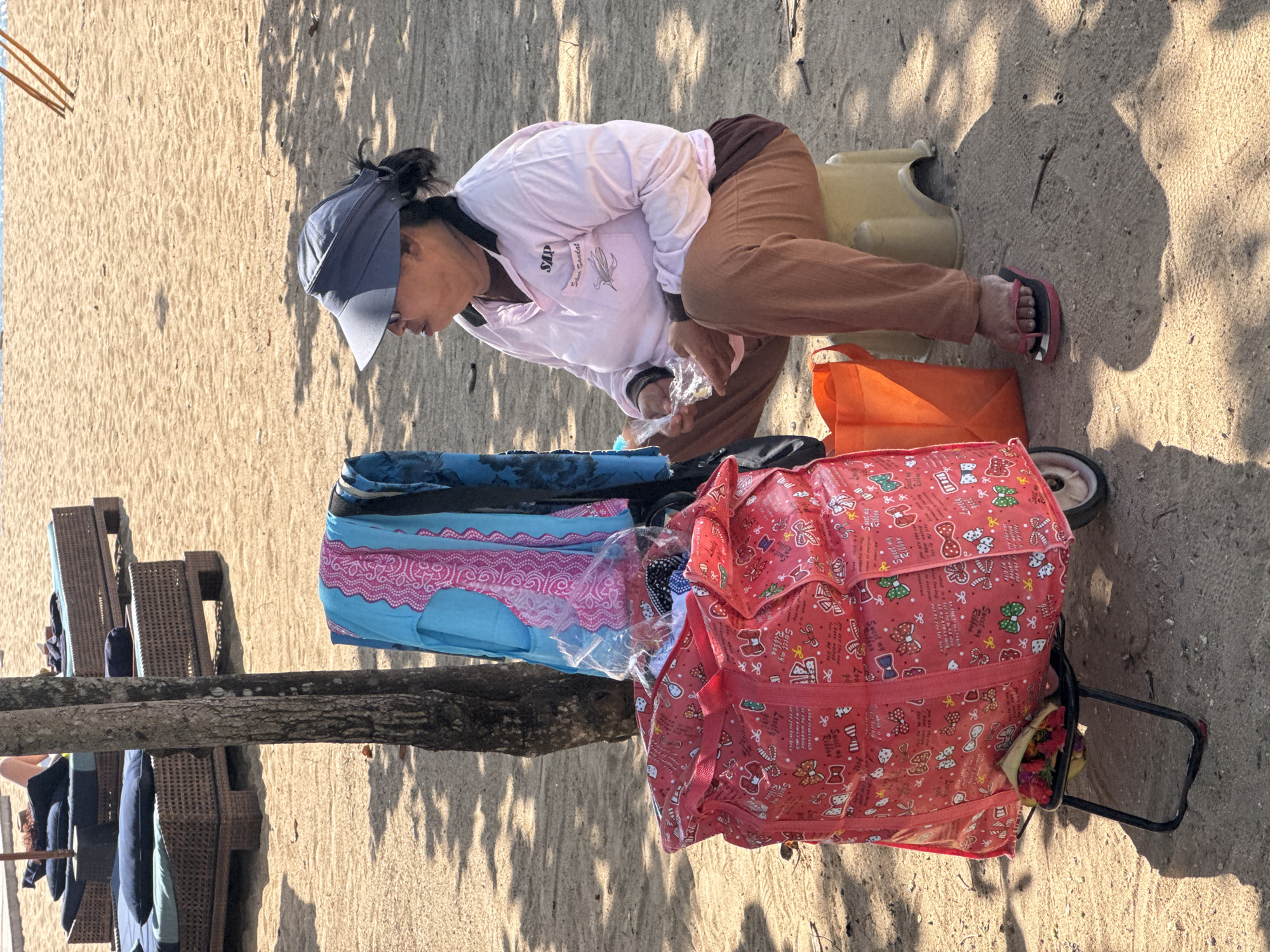 On a sandy surface in Kuta Selatan, Indonesia, a person wearing a blue visor, a light pink long-sleeved shirt with an emblem, and brown pants, is crouching over a display of merchandise. The person is wearing sandals. Beside the person is a small, tan, plastic stool. The merchandise includes items in a blue bag with pink trim, an orange bag, and a large pink patterned tote on wheels. The merchandise appears to be packaged in clear plastic. Nearby are wicker chairs and tables under the shade of a tree.