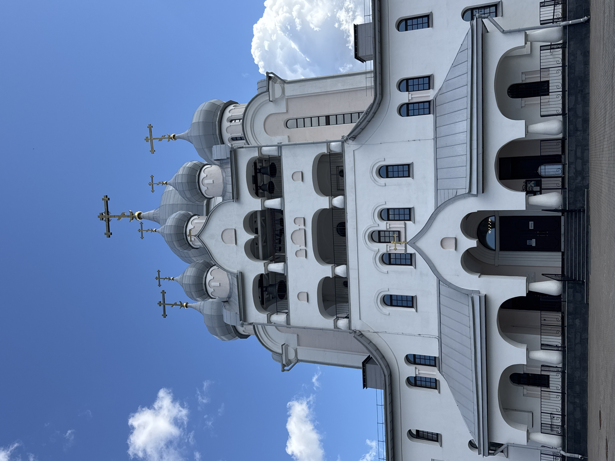 The image depicts the front facade of the Holy Trinity Cathedral of Alexander Nevsky Lavra in Saint Petersburg, Russia. The cathedral is a prominent landmark with multiple domes topped with crosses. The building is predominantly white with grey domes and accents of light pink. The foreground shows a paved area with steps leading up to arched entrances. Several arched windows are visible on the facade, as well as a bell tower with arches and bells. The sky is blue with scattered white clouds. There are no people or any obvious actions or interactions depicted in the image.