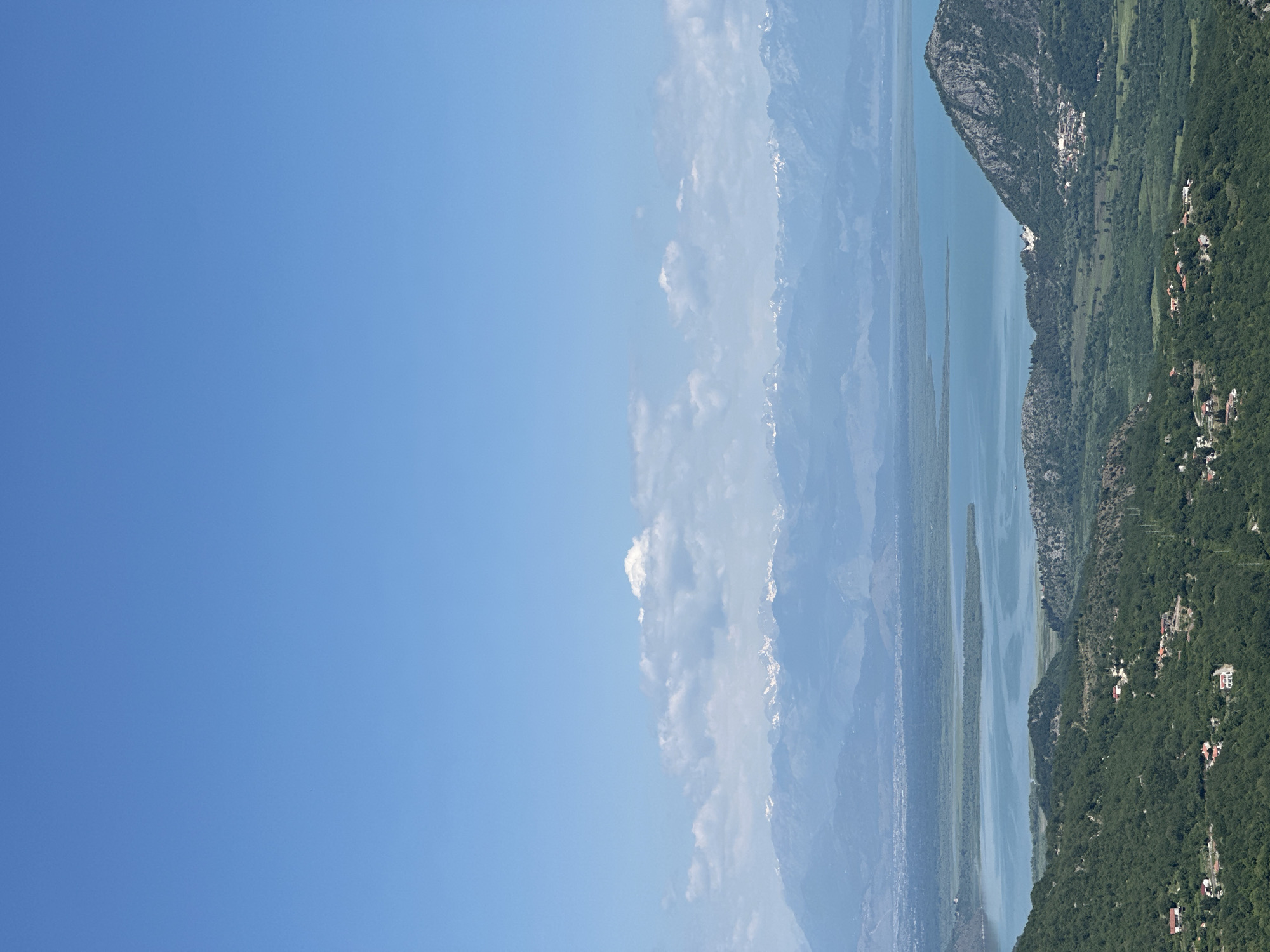 A vertical-oriented scenic shot reveals Bukovik, Montenegro, nestled in a landscape dominated by a tranquil body of water and verdant mountains. A clear blue sky takes up a significant portion of the left frame. Progressing from the left towards the right, a mountain range emerges, some peaks capped with snow. Adjacent to the mountains is a body of water, reflecting the sky. Further right, a densely forested area flanks the body of water, with scattered dwellings.
