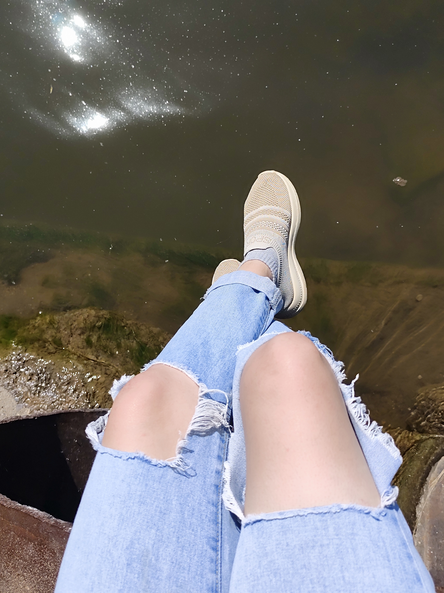 The image is a first-person perspective shot of someone sitting with their legs dangling over what appears to be a body of water. The person is wearing light blue, ripped jeans and light beige slip-on shoes with socks. The water is murky and reflects sunlight. The shore is visible, and appears to be made of rocky and muddy material. The exact location in Фоки, Russia, cannot be determined.