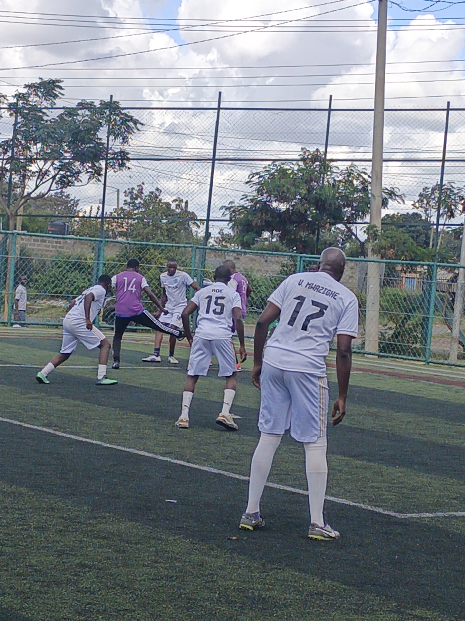 The image captures a soccer match in progress, likely an informal game, on an artificial turf field in Nairobi, Kenya. Six players are visible, dressed in sports attire, with team colors being white and purple. One player wears a white jersey with the number "17" and the name "U. MHAZIGHE." Another sports a white jersey with the number "15" and the letters "ROE". A player in a purple jersey with the number "14" appears to be in motion. Fencing surrounds the field, with trees and buildings visible in the background. A light pole stands near the field's perimeter, and the sky above is partially cloudy.