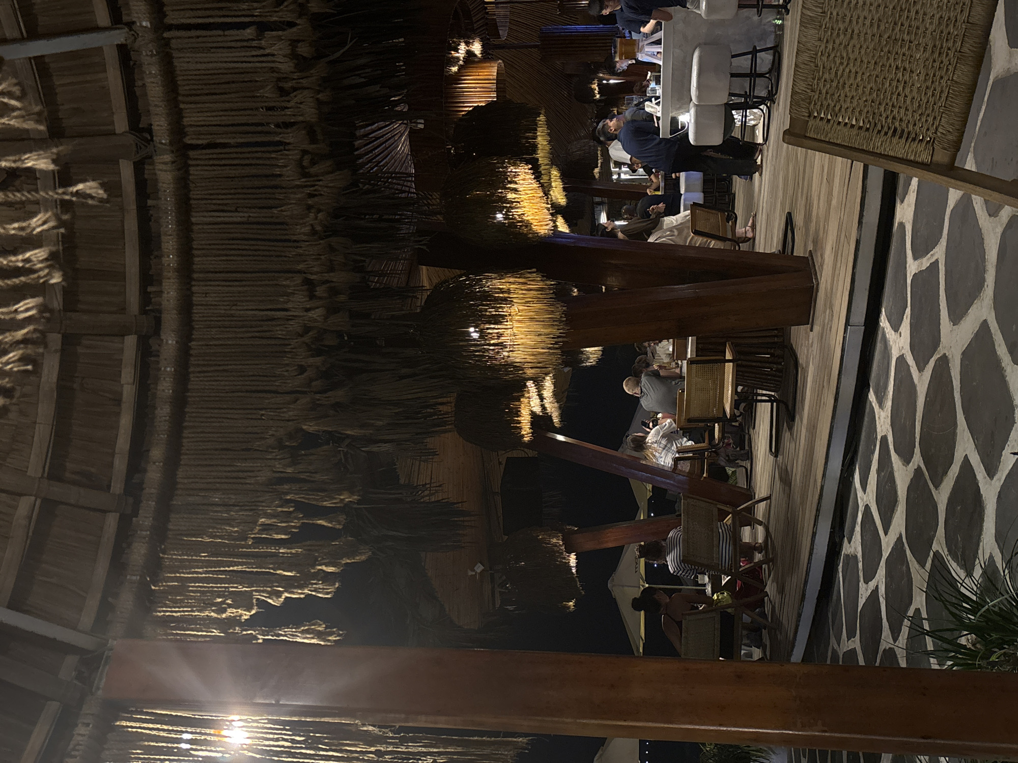 The image depicts an outdoor dining area in Đà Nẵng, Vietnam at night. The location appears to be a restaurant or bar with a thatched roof structure, supported by wooden beams. Patrons are seated at tables and at a bar with white stools. Some people are standing. The flooring consists of wooden planks and a patterned stone or tile design. A woven chair is partially visible. The lighting is dim, with illuminated areas highlighting the dining activity.