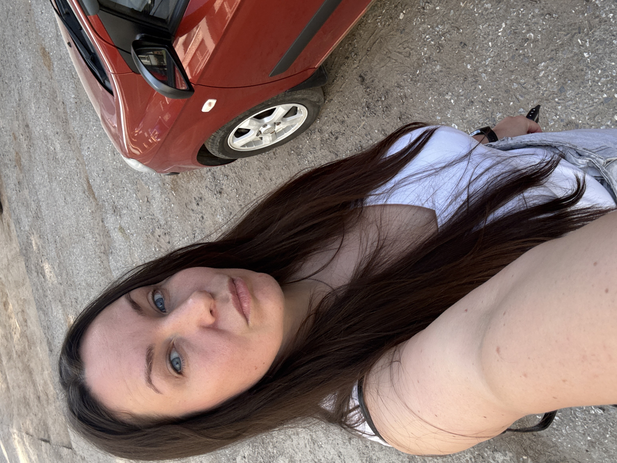 A selfie taken by a person with blue eyes and long, dark brown hair, wearing a white t-shirt, outdoors on a gray concrete surface. The person's left arm is visible, with a black watch on the wrist. A red car is partially visible in the top left corner of the frame. The location appears to be an outdoor parking area or street. The location is Chaikovsky, Russia, but there is no information in the image to verify this.