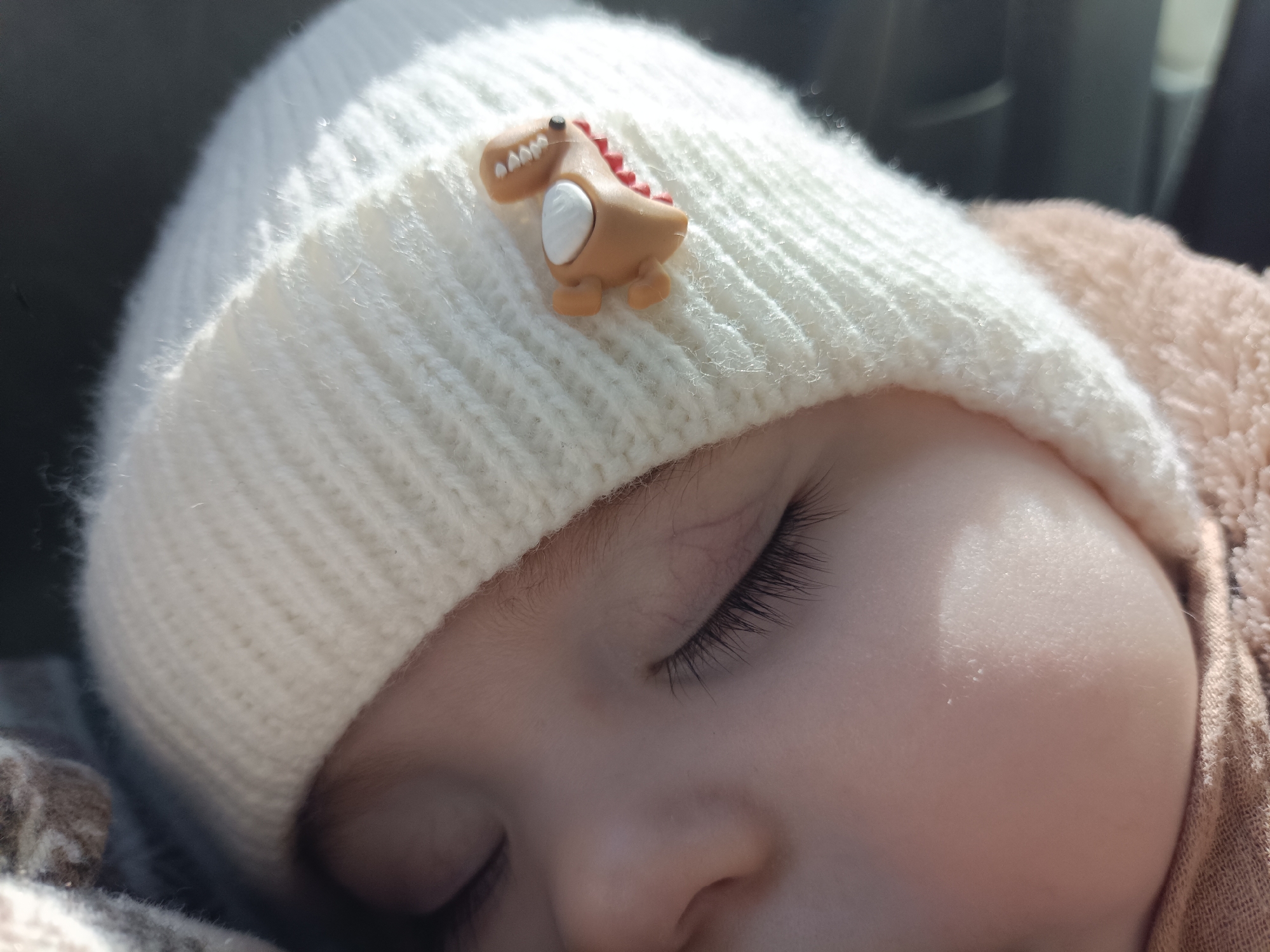 The image displays a close-up, high-angle view of an infant or young child's head, located in the city of Chaikovsky, Russia. The child is wearing a white, ribbed knitted beanie adorned with a small, light brown dinosaur appliqué positioned towards the front. The dinosaur figure features white teeth and red spikes along its back. The child's eyes are closed, indicating sleep, with distinct dark eyelashes visible. Portions of the child's nose and cheek are also present in the frame. A light brown or peach-colored textured fabric, possibly part of clothing or a blanket, is visible along the child's right side, extending from beneath the beanie. A darker, patterned fabric is visible on the lower left. Strong directional light illuminates the upper left portion of the beanie and the child's skin. The background is dark and blurred, suggesting an indoor or enclosed environment.