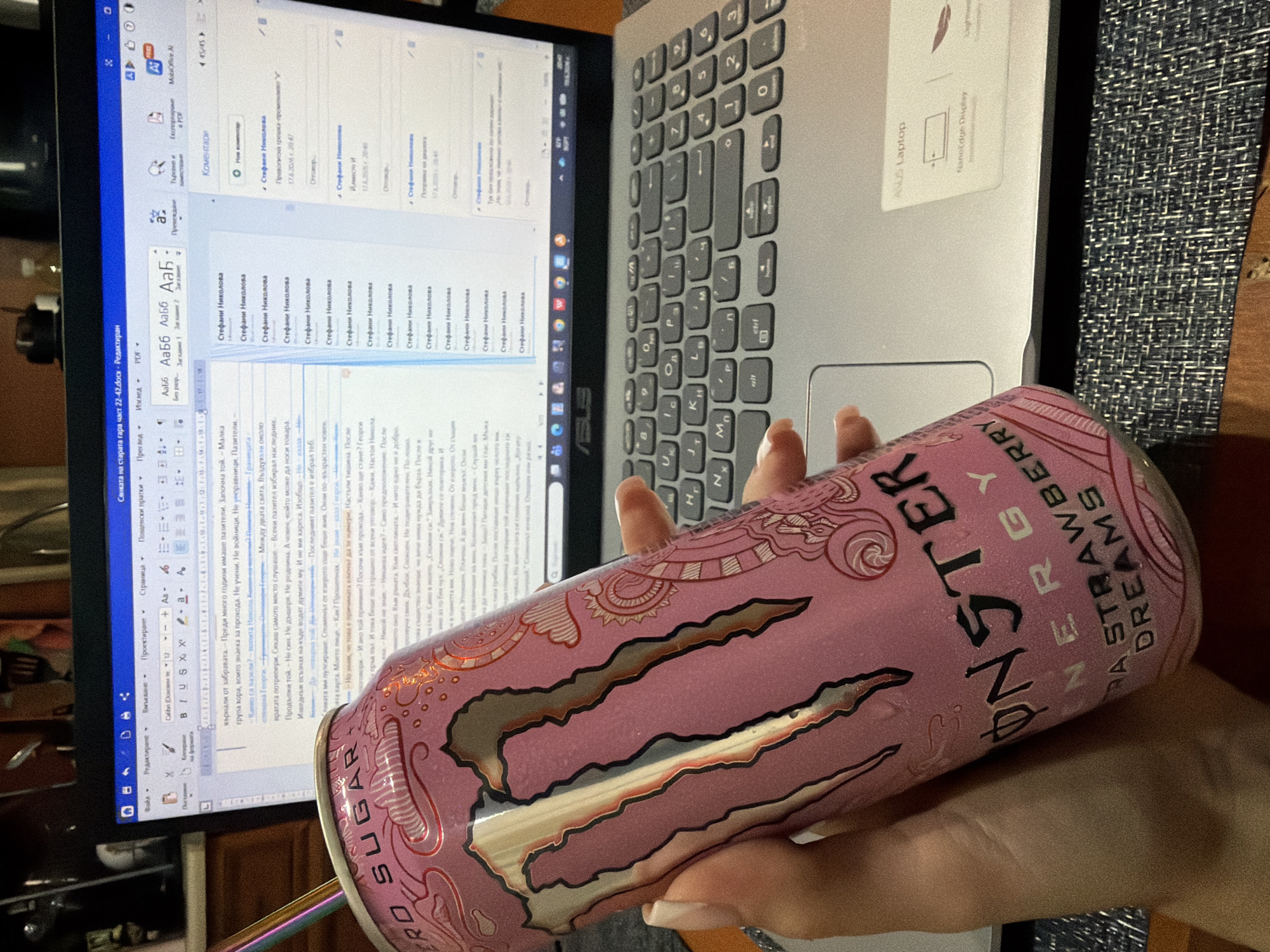 The image depicts a close-up view of a pink Monster Energy Ultra Strawberry Dreams can held by a person's hand in the foreground. In the background, a laptop is open and displays a document with text in Bulgarian. The document appears to be a word processing file, possibly with comments or tracked changes visible. The laptop is placed on a desk or table, and part of the keyboard is visible. The overall setting suggests a casual workspace, possibly at home. There is no specific visual information in the image that would allow for the determination of a location within the city of Burgas, Bulgaria.