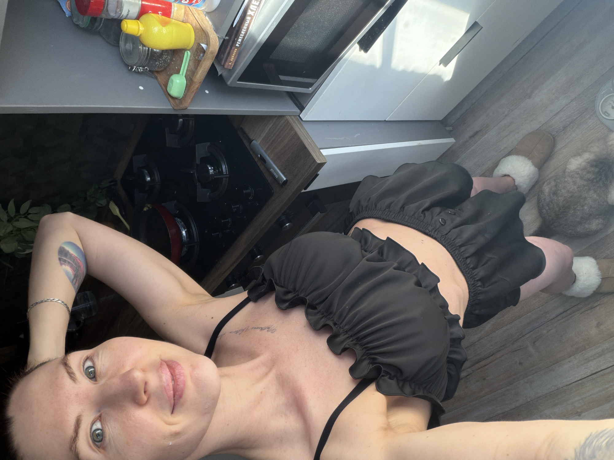 A young, Caucasian woman is lying on a wooden floor in a kitchen, seemingly taking a selfie. She is wearing a black ruffled bra-style top and matching shorts. Her left arm is raised above her head, displaying a tattoo. She is wearing a silver bracelet on her left wrist. Her hair is pulled back, and she looks directly at the camera with a slight smile.
The kitchen features grey countertops with various items including spices, a yellow bottle, and cooking utensils. A black glass-top stove is below the counter. A microwave oven is positioned above the stove. White cabinets are partially visible in the background. She appears to be wearing fur-lined slippers. A grey cat sits behind her.
It is not possible to determine if this image was taken in Perm, Russia, based on the provided information.