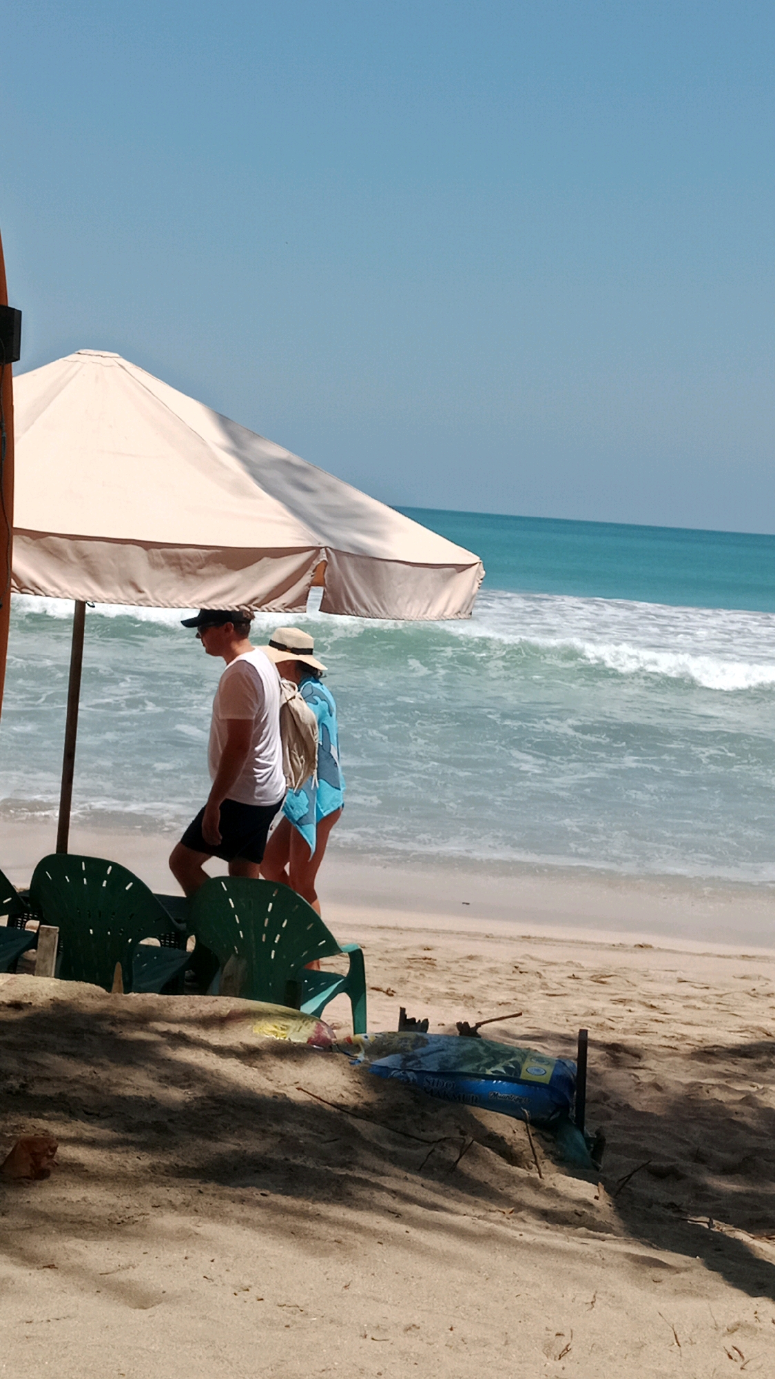 The image depicts a beach scene in Kuta, Indonesia, under a clear blue sky. A large, beige beach umbrella shades two people walking on the sand, with the ocean waves breaking in the background. The man on the left wears a white t-shirt, black shorts, and a dark baseball cap. The woman beside him has on a straw hat and a blue and white patterned cover-up. Two green plastic chairs are visible in the foreground, partially obscured by shadows cast from the umbrella and nearby foliage. On the sand in the lower right portion of the image, a blue bag with writing on it lies next to some debris. The ocean is a vibrant turquoise, with white foam on the crests of the waves.