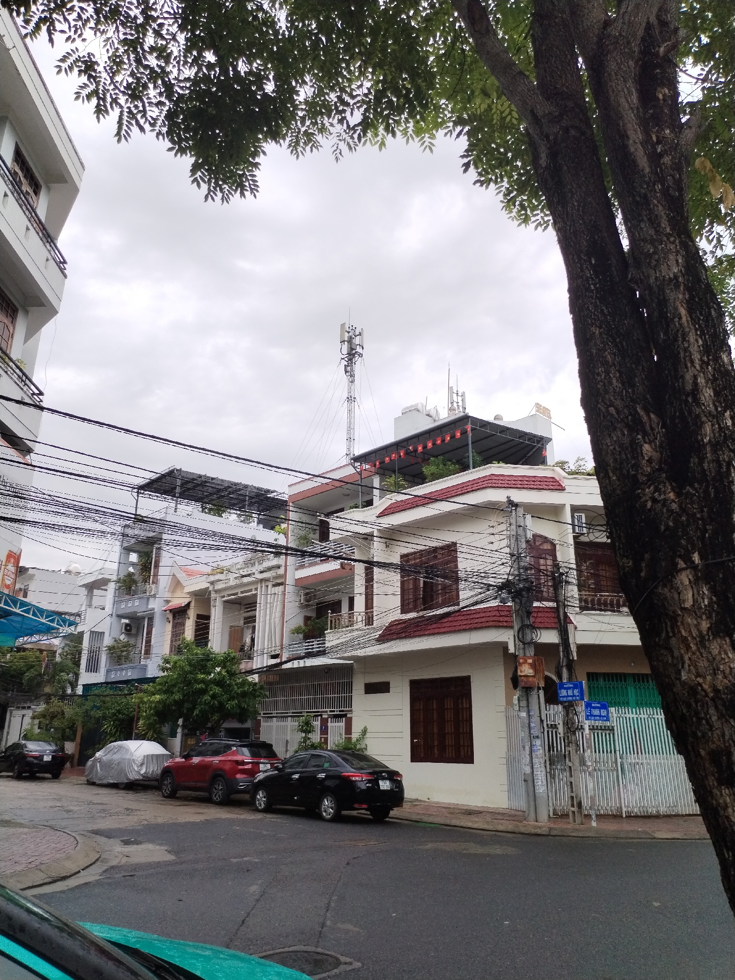 This media file captures a street view within a residential area of Nha Trang Ward, Vietnam, under an overcast sky. The scene features a row of multi-story buildings, a street with parked cars, and a dense network of overhead wires.
The buildings are primarily white with red or brown tiled roofs, featuring balconies with metal railings, and windows often fitted with wooden shutters or ornate bars. On the far left, a modern white building with dark-framed windows is partially visible. Towards the center-right, a rooftop is adorned with a structure that appears to be solar panels or a canopy, from which small Vietnamese flags are hung. On a rooftop further in the background, a tall cellular tower with multiple antennas is prominent.
On the asphalt street, several vehicles are parked: a black sedan on the far left, a car covered with a light-colored tarp, a red SUV, and another black sedan. The front part of a green vehicle is visible in the bottom left corner. A tiled sidewalk runs alongside the street, bordered by white fences and gates in front of the houses.
Numerous black electrical and communication wires crisscross the sky, connecting to utility poles that feature various attached boxes and signs. A large tree with a dark, textured trunk stands prominently on the right side of the frame, with its green foliage extending overhead, partially obscuring the sky.
A notable detail is a blue street sign attached to a utility pole on the right side of the street. It displays two lines of text: "LƯƠNG NGU HỌC" at the top and "LÊ THANH NGHỊ" below, indicating street names. The time of day appears to be daytime, though the heavy cloud cover suggests diffused light rather than bright sunshine. No people are visible, and the scene conveys a calm, ordinary urban atmosphere.