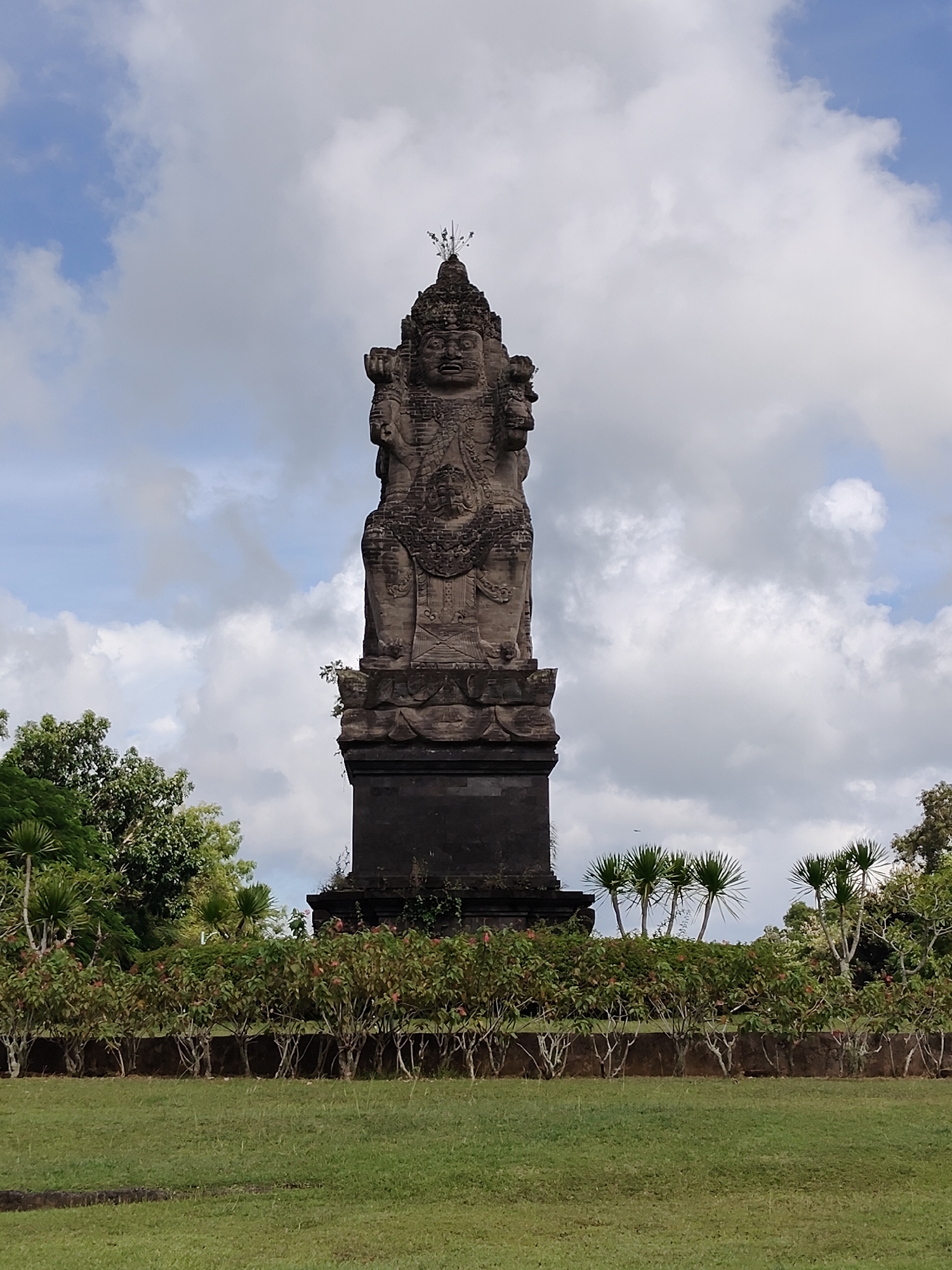 The image shows a tall, weathered stone statue with multiple faces, likely a deity or a mythical figure, standing on a tiered base in Pecatu, Indonesia. The statue is positioned in a grassy area with a low wall and a row of small bushes in front. Trees surround the area. The sky is cloudy with patches of blue. No people are visible. The scene appears to be a public park or garden setting.