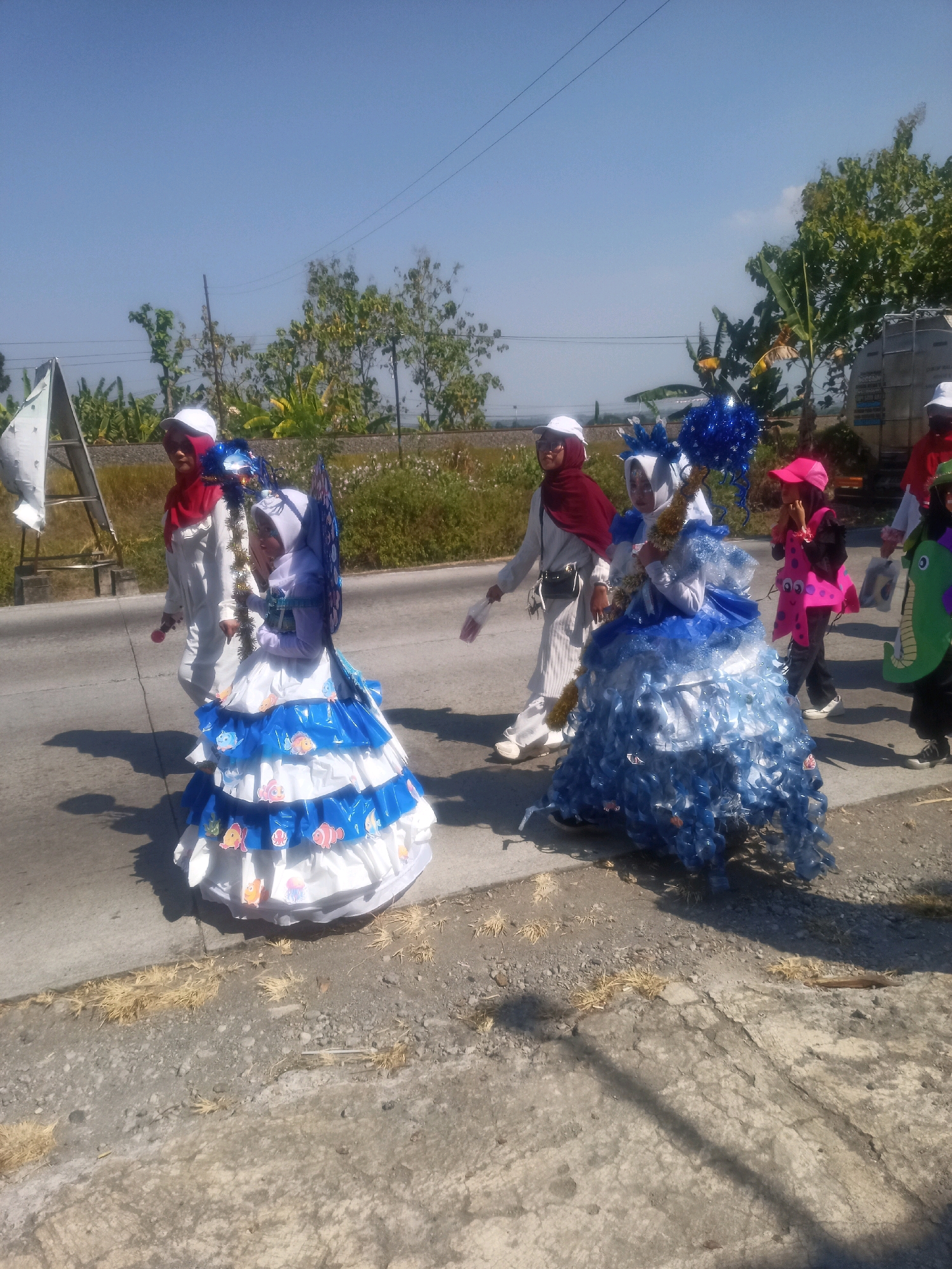 In Sumberlawang, Indonesia, a group of children in festive costumes walks along a paved road under a clear blue sky. The central figures are two girls dressed in elaborate outfits. The girl on the left wears a white dress adorned with blue and white ruffles, decorated with colorful fish cutouts. Her dress has a voluminous, tiered skirt, and she wears a white hijab with a red shawl. She is holding a large, blue, spiky headdress on a stick. To her right, another girl is dressed in a bright pink costume shaped like a cartoon character, possibly a monster or creature, with large eyes and a wide smile. She wears a pink hat and dark pants.
Other individuals are present in the procession. To the left of the two main figures, a person in a white outfit and a white cap walks with a red object in their hand. Further to the right, a person in a green costume resembling a dinosaur or sea creature is visible. The background consists of lush green vegetation, trees, and a distant, partially obscured truck. The road has a dry, grassy verge. The overall scene depicts a lively street event or parade, likely a cultural or festive occasion.