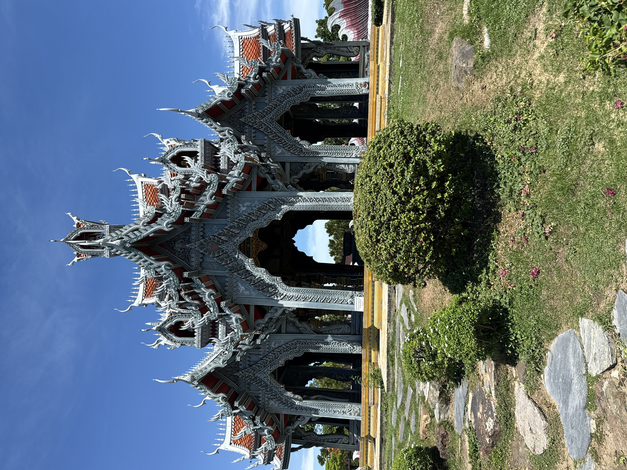 The image shows an ornate, multi-tiered pavilion structure with a gray facade and red roof accents. It is surrounded by meticulously maintained green lawns and stone pathways. The location is Bang Pu Subdistrict Municipality, Thailand. A rounded, neatly trimmed bush is positioned to the right of the structure. The sky is clear and blue.
