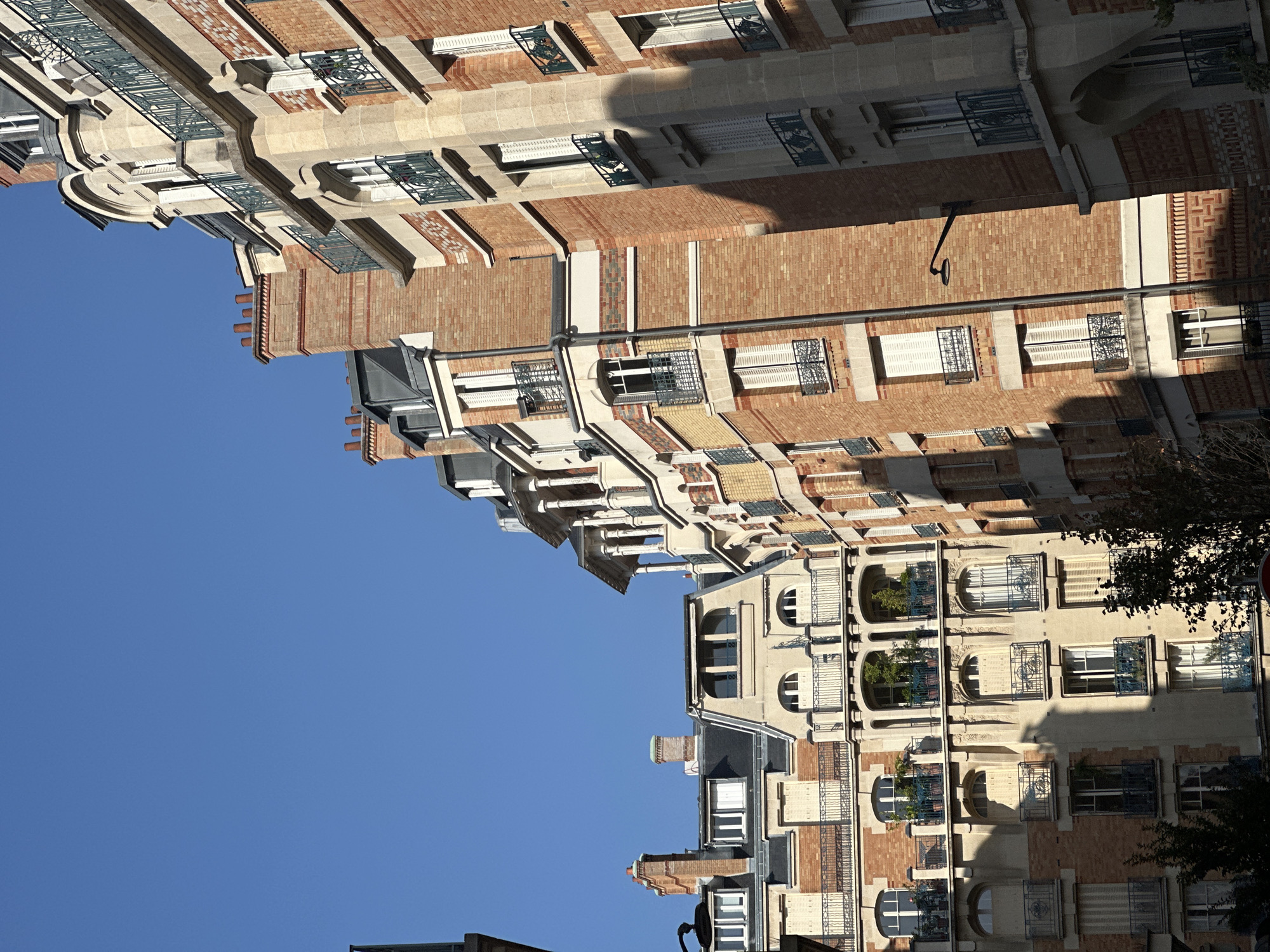 This image shows a series of residential buildings in Paris, France, under a clear blue sky. The buildings are constructed with a mix of red brick and light-colored stone, featuring classic Parisian architectural elements such as mansard roofs, balconies with ornate railings, arched windows, and dormer windows. The facades are decorated with intricate brickwork patterns and stone detailing. Chimneys with decorative tops are visible on the rooftops. Some balconies are adorned with potted plants, adding touches of greenery. The sunlight casts shadows on the buildings, highlighting their architectural features and creating a sense of depth. There are no people visible in the image. The scene appears to be a typical residential street in a Parisian neighborhood.