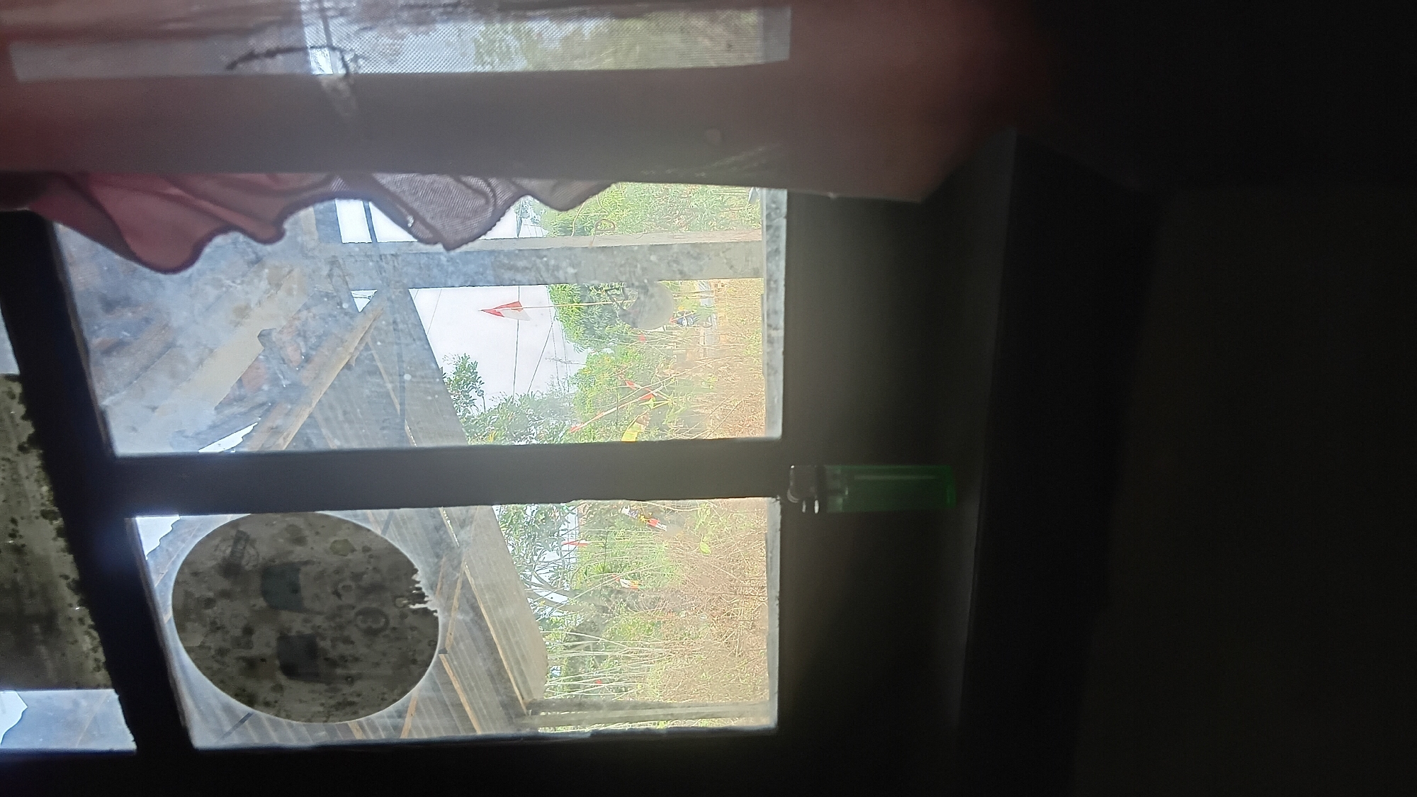 A close-up shot captures a grimy windowpane looking out onto an outdoor scene. The window is divided into two panes by a dark vertical frame. A circular sticker, possibly depicting a moon with two dark squares inside, is adhered to the left pane. A green plastic lighter rests on the dark ledge below the window.
The view through the window reveals an overgrown area with tall, dry grass and various trees in the background. Several small flags, predominantly red and white, are scattered throughout the landscape, suggesting a festive or commemorative occasion. The sky is overcast. To the right side of the frame, a portion of a pinkish-red curtain is visible, partially obscuring the view. The overall environment suggests a rural or semi-rural setting, possibly within or on the outskirts of Prabumulih, Indonesia, given the presence of Indonesian flags. The conditions of the window and the surrounding area imply a state of disrepair or neglect.