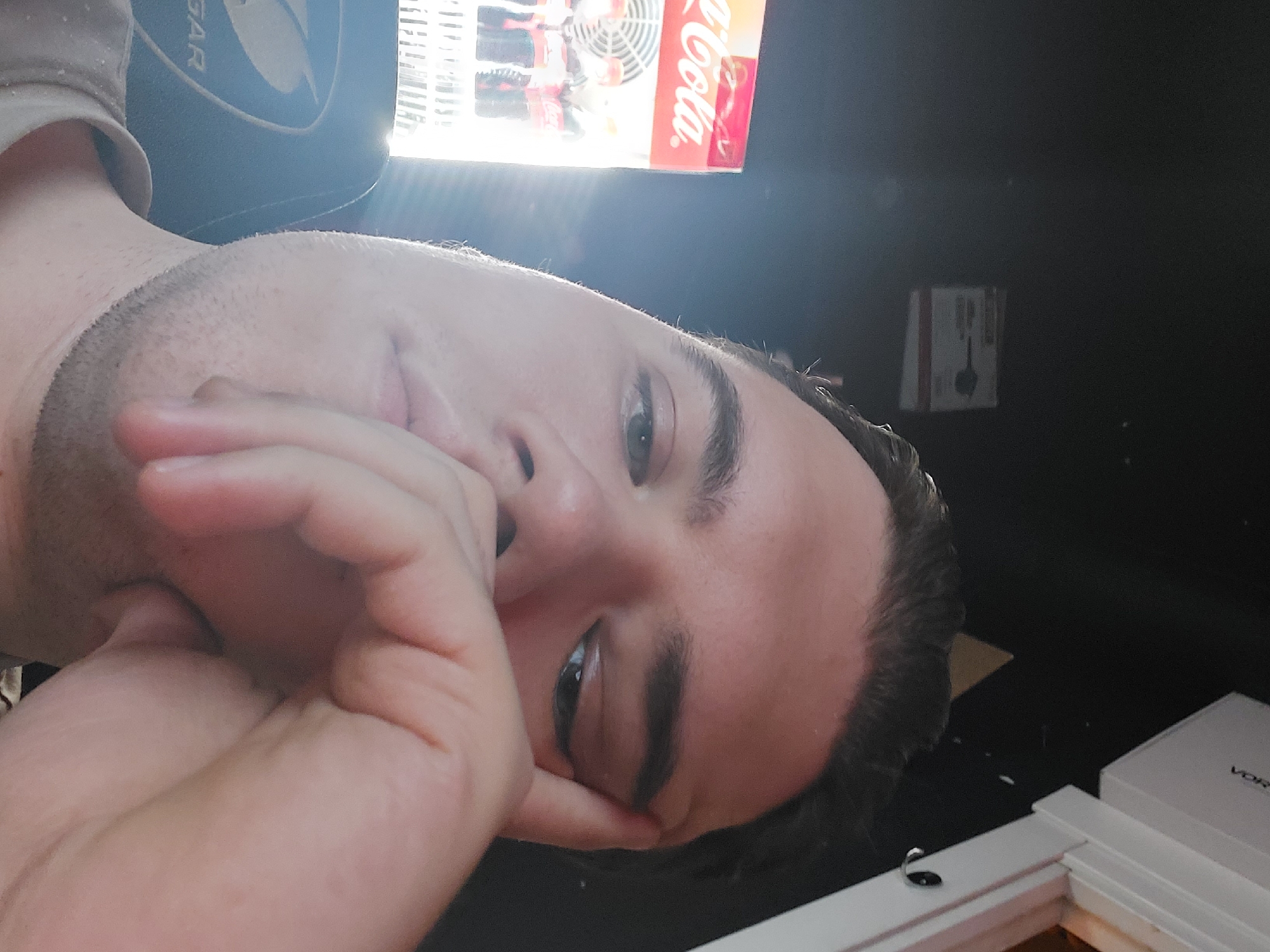 A man with short, dark hair and light skin is resting his face on his hand. The photo is taken at a close, angled perspective. Above his head is a cooler, advertising Coca-Cola. In the background, there's a dark wall and what appears to be a box hanging on it. In the lower right corner of the image, there is a white box on a white surface. This image was not taken in Tashkent, Uzbekistan.