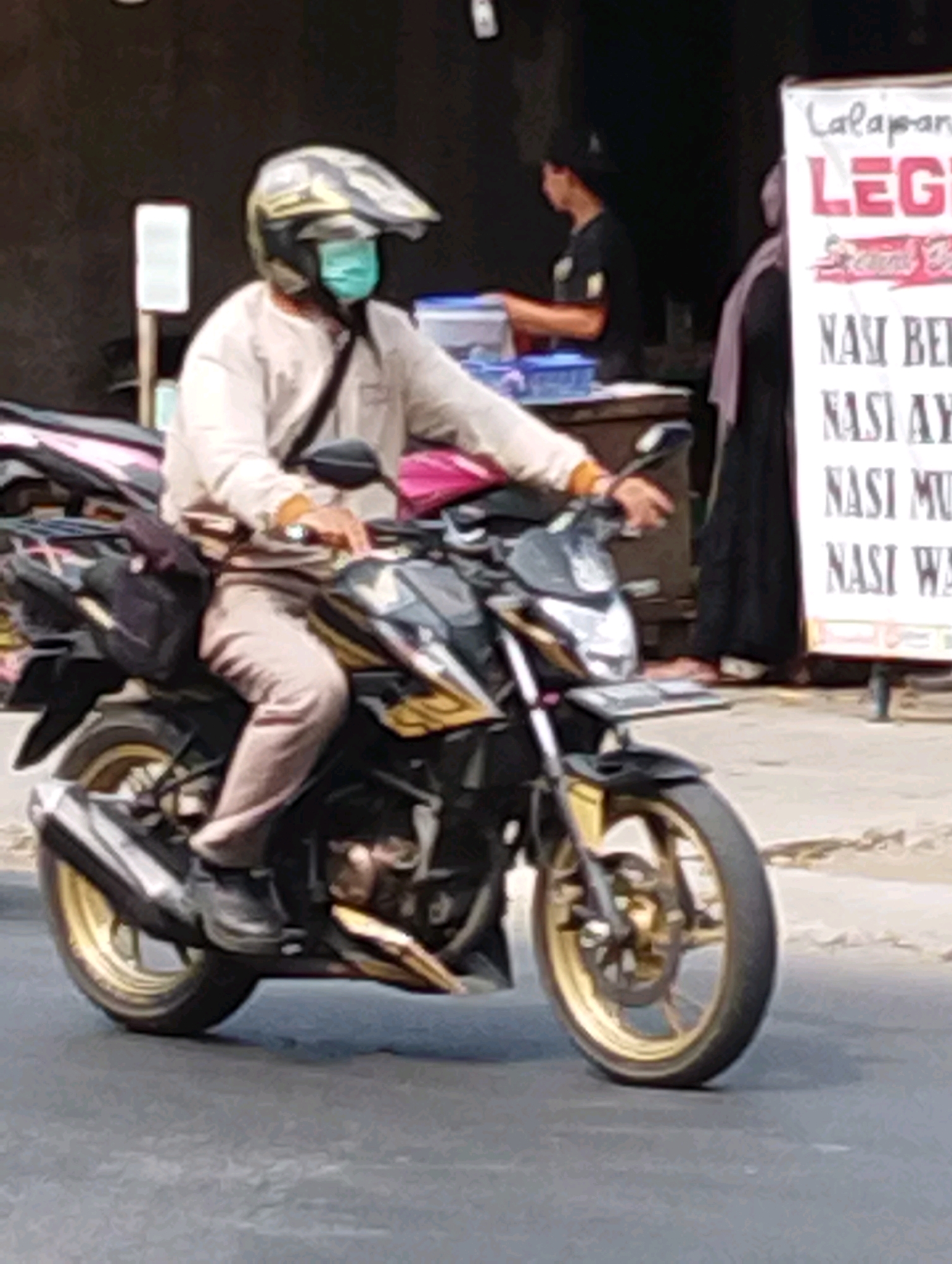 A person wearing a helmet and a face mask rides a motorcycle in Manaruwi, Indonesia. The person is wearing a light-colored long-sleeved shirt and light-colored pants. The motorcycle is black and gold with a silver exhaust pipe. In the background, there is a sign with Indonesian writing, indicating a food establishment. There are other people and a stall visible behind the rider. The scene appears to be on a street.