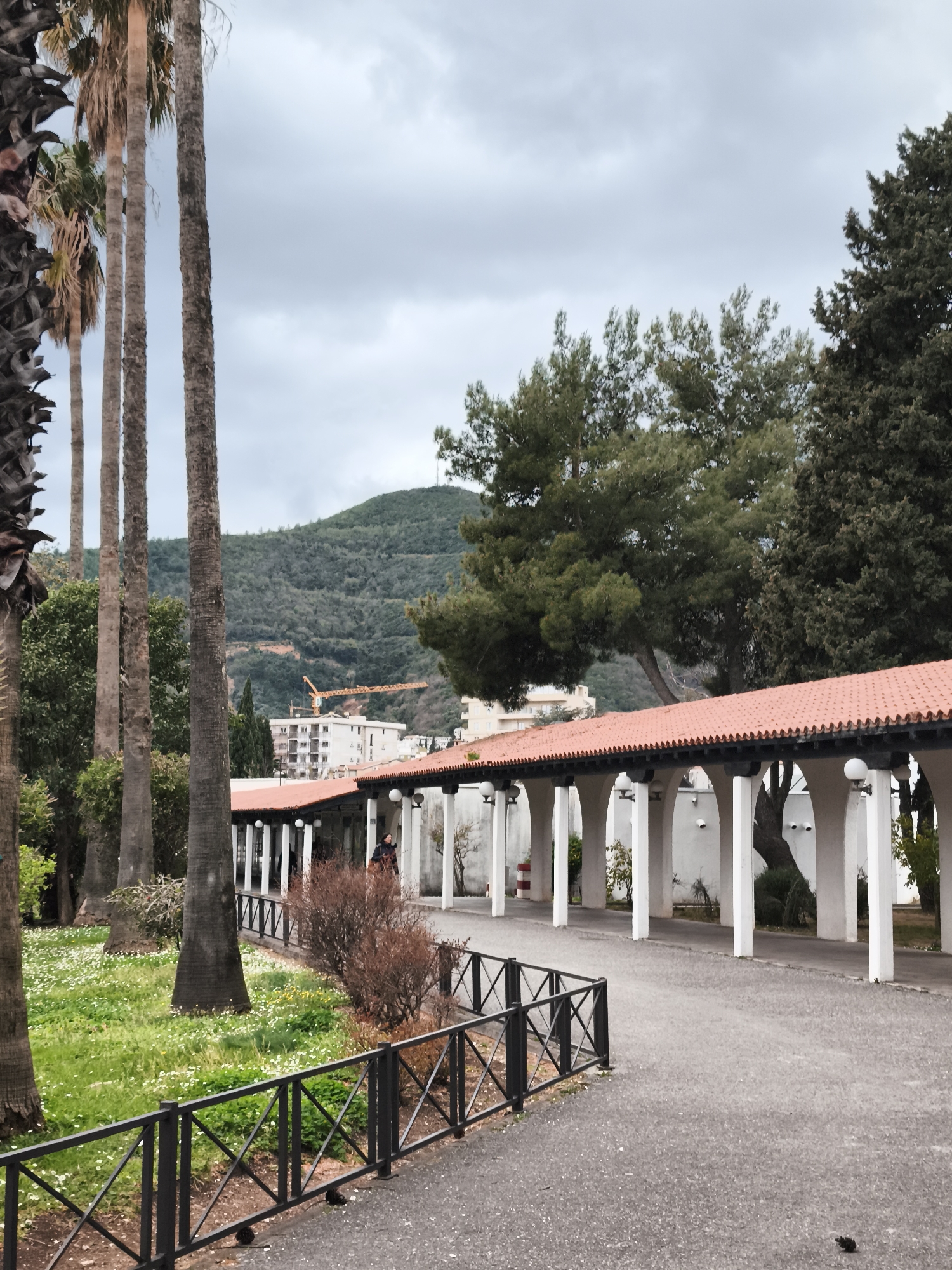The image shows a park scene with a walkway lined by a colonnade, likely taken in the city of Budva, Montenegro. The colonnade has a red-tiled roof and white arched supports with spherical lamps attached to the structure. Behind the colonnade, there is a white building. In the background, a green mountain range is visible under an overcast sky. In the foreground, a black metal fence runs along the walkway. Palm trees are present on the left side of the image, and various other trees and bushes are scattered throughout the park area. The ground is covered with grass and small white flowers in the park area, while the walkway is paved. There appears to be a person walking under the colonnade. A construction crane can also be seen in the background.