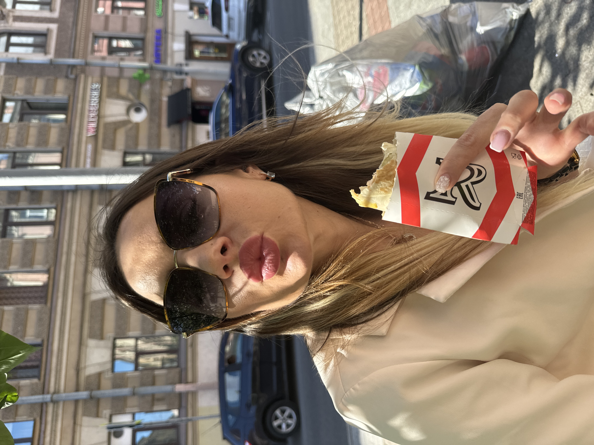 A woman is taking a selfie outdoors, holding a half-eaten food item wrapped in paper. She is wearing large sunglasses, a beige blazer, and a necklace. Her lips are puckered, and her hair is long and blonde. In the background, a street scene in Saint Petersburg, Russia, is visible, with buildings, cars, and a tree branch in the foreground. One building has a sign that reads "WILDBERRIES".