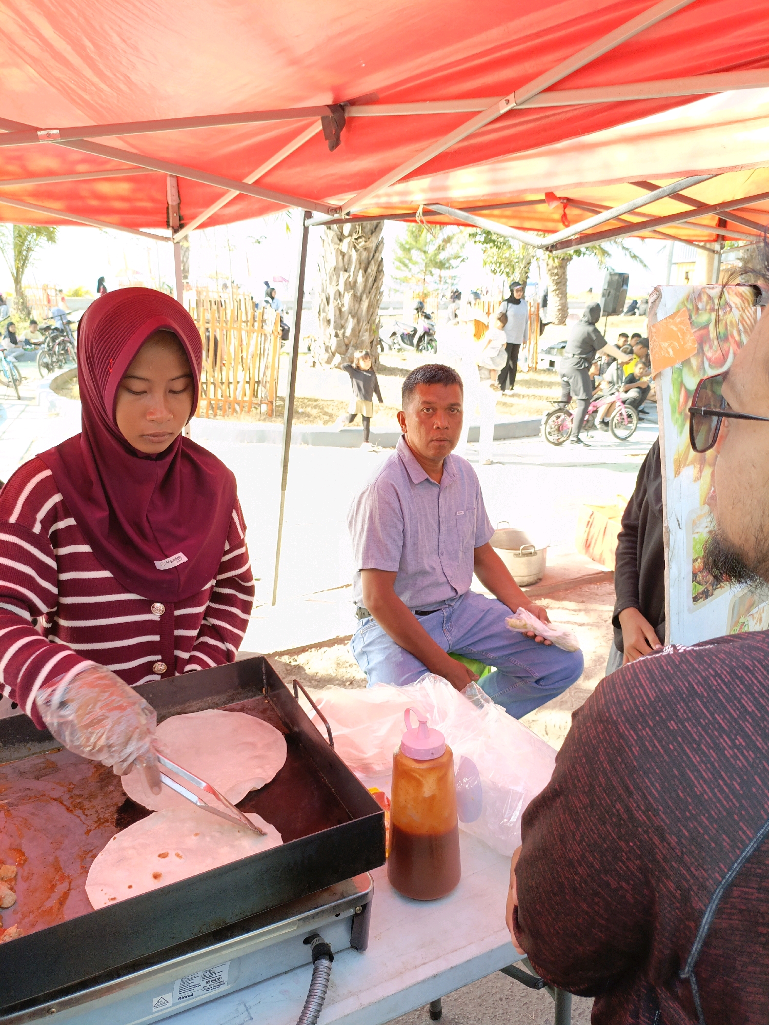 In Bulukumba, Indonesia, under a bright red canopy, a street food vendor is preparing flatbread on a griddle. A young woman, wearing a maroon hijab and a striped sweater, uses tongs to turn the flatbread. Beside her, a man in a light blue collared shirt and jeans sits on a chair, holding a plastic bag of food. Other people and activities are visible in the background, suggesting a bustling outdoor market or gathering. The scene is illuminated by natural sunlight.