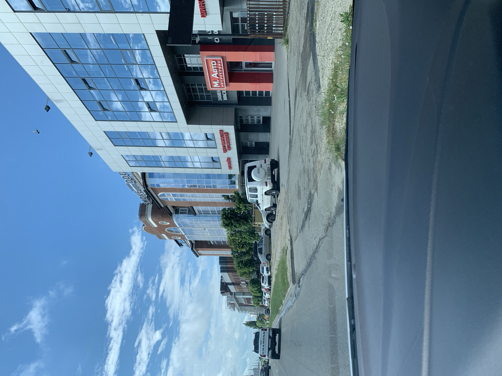 The image captures a view from a vehicle driving down a street in Stavropol, Russia, on a partly cloudy day. The street is lined with modern, multi-story commercial buildings. The building on the right features a prominent red and white sign that reads "М. АВТО АВТОСЕРВИС," indicating an auto service center. Adjacent to this is a building with a brick facade and large arched windows. Further down the street, other buildings with glass facades and signage are visible. Several cars are parked along the roadside and in the street, including a white SUV and a white pickup truck with "CHANGAN" written on its tailgate. The foreground of the image shows the interior of the vehicle, including the dashboard. The environment appears to be a commercial district with clear signage and a mix of architectural styles.
