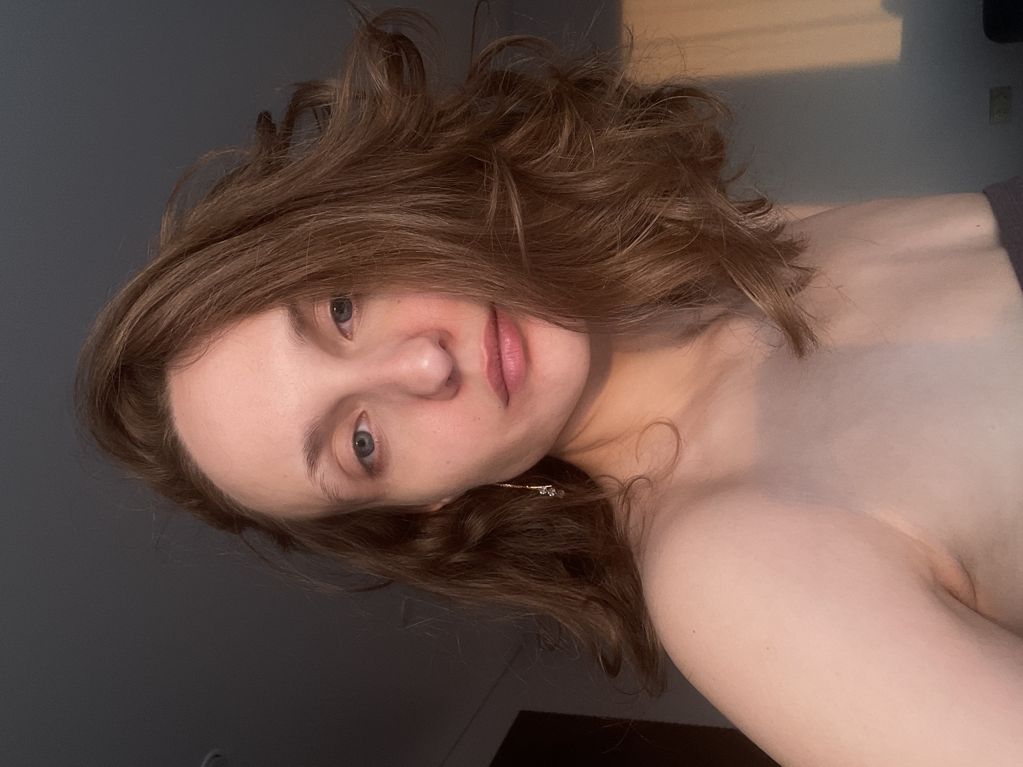 A close-up selfie captures a person, likely female, with medium-length, wavy brown hair, light skin, and blue eyes. The person is looking directly into the camera with a neutral expression, their head slightly tilted. The left side of the image features a plain dark grey background, while the right side shows a lighter, possibly off-white or tan wall, illuminated by a warm, directional light source that casts subtle shadows. A golden dangle earring with a clear stone is visible near the person's left ear. The person's bare shoulders and upper chest are visible, suggesting they are wearing a top with a wide neckline or are shirtless, with a dark grey fabric visible on the far right edge of the frame, possibly a garment or towel. The lighting suggests an indoor setting, likely late afternoon or evening, given the soft, warm glow. There is no visible text. The provided location context is Podshivalovo, Russia.
