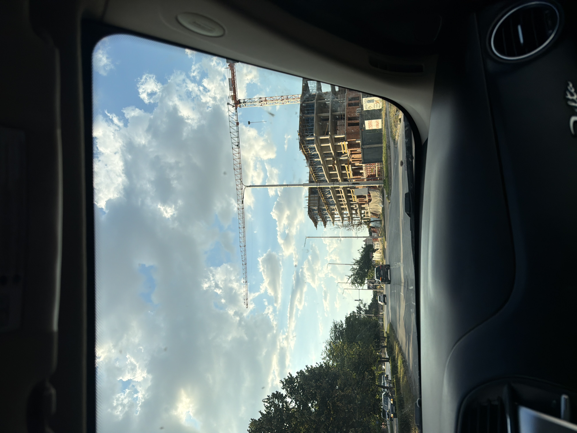 This image captures a view from inside a vehicle looking out at a street scene in Georgi-Dobrevo, Bulgaria. The perspective is that of a passenger or driver, with the dashboard and windshield frame visible in the foreground.
The outdoor scene is dominated by a bright, partly cloudy sky with dramatic cumulus clouds. A long construction crane, typical of urban development, stretches horizontally across the middle ground. To the right, a multi-story building under construction is visible, showing concrete framing and some brickwork.
The street itself is paved and appears to be a main thoroughfare with multiple lanes. Several cars are driving on the road, including a dark SUV in the right lane and other vehicles further down. Trees line the left side of the street, providing a green contrast to the construction. Streetlights and traffic signals are also present.
The overall impression is one of an active urban area undergoing development, under a dynamic sky.