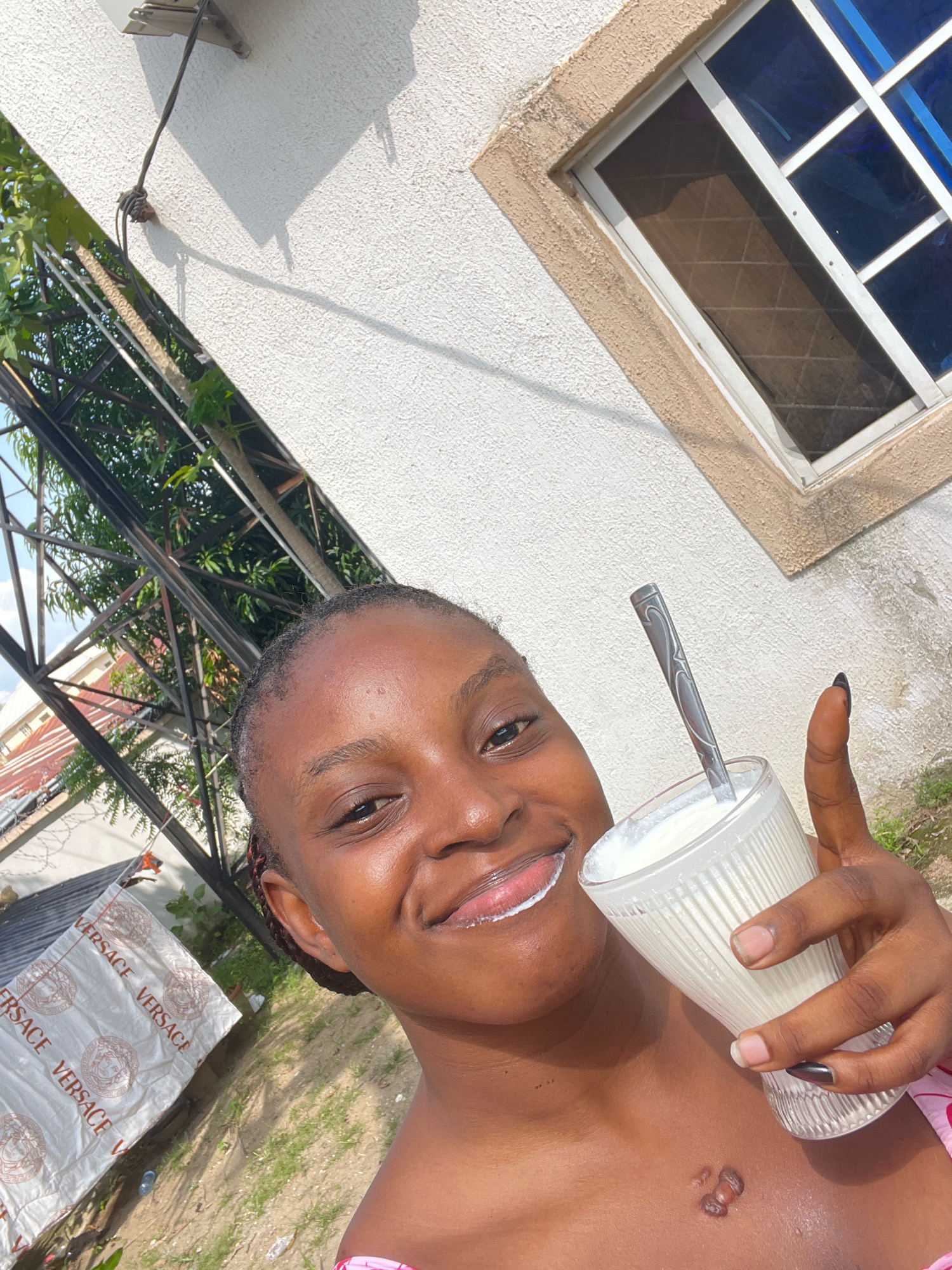 A young Black woman is taking a selfie outdoors in front of a textured, white wall and a blue-paned window. She is holding a glass of white liquid with a spoon in it and smiling at the camera, with a bit of the liquid on her lip. She has a mark on her chest. In the background, there is green foliage, a metal structure, and a rolled-up white sheet with the word "VERSACE" printed on it. The ground is covered with sand and grass.