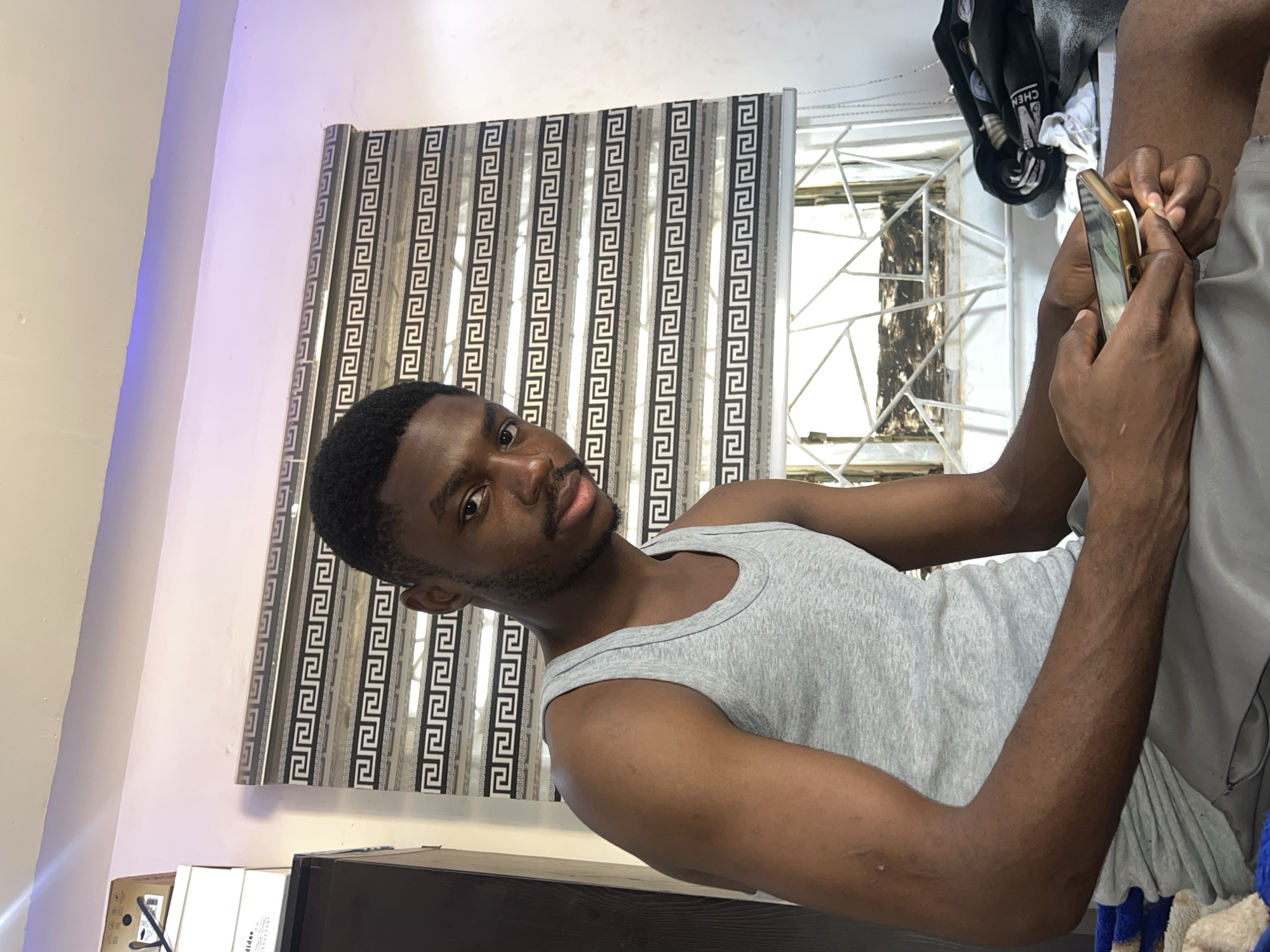 The image, possibly captured in Jos, Nigeria, features a young man of African descent looking directly at the camera with a neutral expression. He is wearing a plain grey tank top. His hands are visible on the right side of the frame, holding a gold-colored smartphone that has a white pop socket attached, suggesting he is interacting with the device.
In the background, a window is partially covered by vertical blinds adorned with a repetitive black and white Greek key (meander) pattern. To the right of the blinds, a window with white security bars is visible. Further in the background, towards the upper right, some folded clothes are stacked, including a black item where the partial text "CHEN" can be seen.
The setting appears to be an indoor room, possibly a private residence. On the left, a light-colored wall features a subtle blue or purple LED strip light near the ceiling. Towards the bottom left, a dark surface, likely furniture, is visible, along with some white objects; one item displays the partially visible text "adidas." The lighting indicates it is daytime, with natural light entering the room, complemented by indoor illumination.