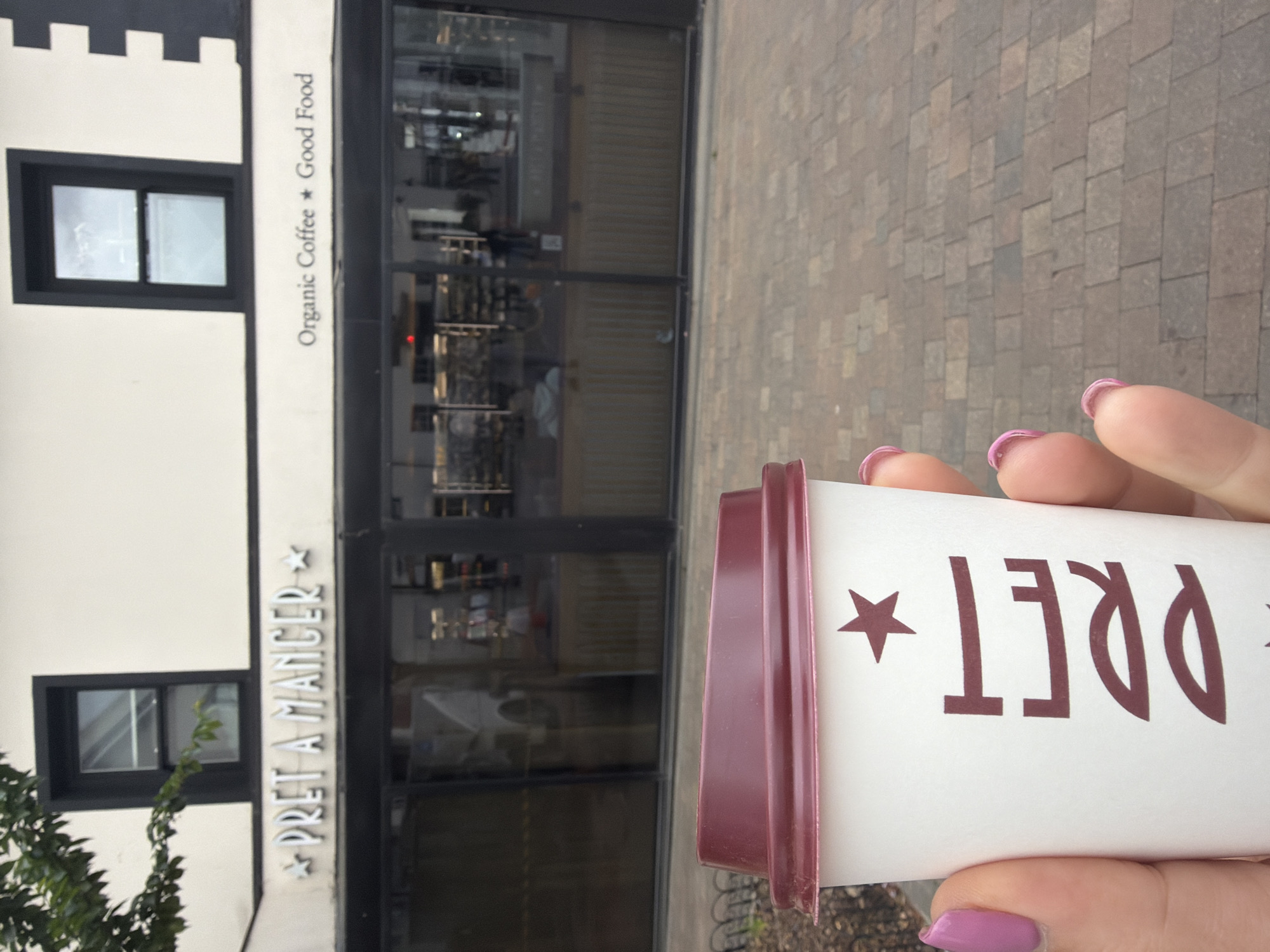 A cup from the Pret A Manger restaurant is being held up in front of the restaurant location in Epsom and Ewell, United Kingdom. The cup is white with the Pret A Manger logo printed in burgundy. The lid of the cup is also burgundy. The person holding the cup is visible from the fingers, with fingernails painted a light purple. The restaurant is a white building with black framing around the windows. The name "Pret A Manger" is printed vertically on the side of the building, as is "Organic Coffee * Good Food". The restaurant has a large glass front with multiple doors. The sidewalk in front of the restaurant is made of brown bricks.