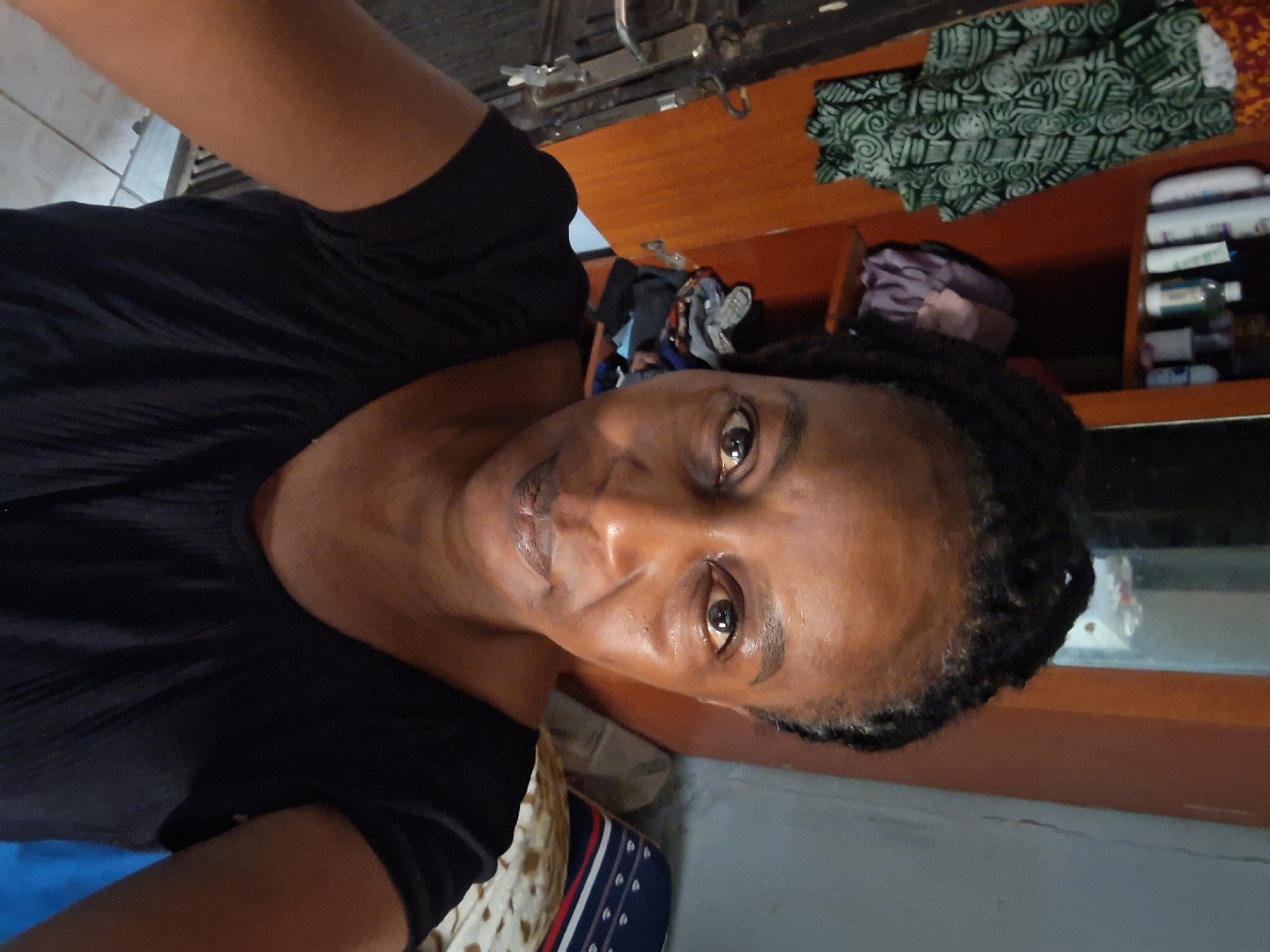 A woman with dark skin and braided hair is the primary subject. She is wearing a black shirt. The background suggests an interior space, potentially a sewing area, given the presence of a sewing machine and fabrics. Other items include shelves with bottles, and a blue surface in the foreground. There is no discernible information in the image that confirms this scene is in Sabon Tasha, Nigeria.
