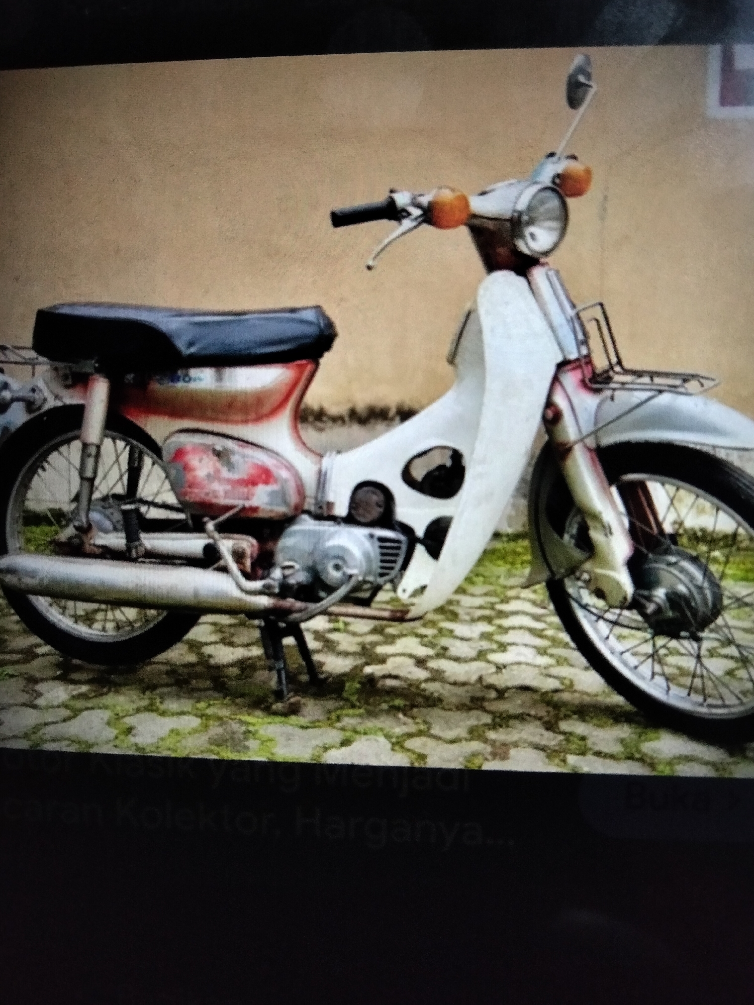 A weathered, red and white vintage motorcycle is parked on a brick-paved surface with patches of moss. The motorcycle has a black seat, a silver headlight with orange turn signals, and a prominent white front fairing. Its chrome exhaust pipe is visible, as are the spokes of the wheels. The background consists of a textured, light brown wall. The scene appears to be outdoors, possibly in a less maintained area, given the condition of the motorcycle and the surrounding ground. The image is taken from a slightly elevated angle. The lower portion of the image is obscured by a dark area with faint, illegible text. The overall impression is that of a static display of a classic vehicle. There is no indication of the city of Kualaraja, Indonesia, in the image itself.