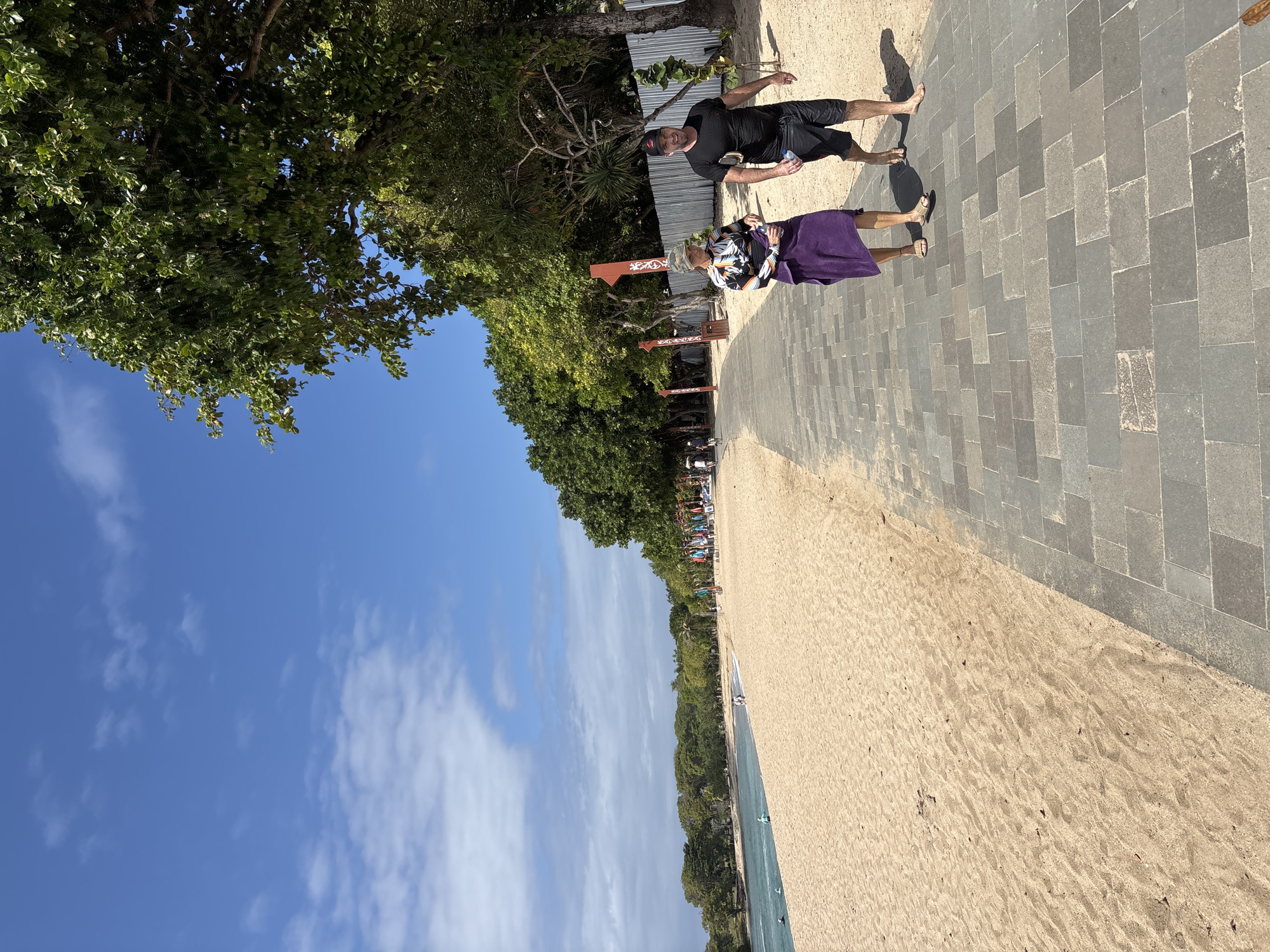 The image depicts a scenic view of a beach in Kuta Selatan, Indonesia. The scene features a sandy beach stretching towards the ocean, bordered by lush green trees and a paved walkway. Two people are visible walking on the paved path. A man in a black t-shirt and shorts walks ahead, while a woman in a colorful top and a purple skirt follows behind. The time of day appears to be afternoon, with the sun casting shadows on the ground. The weather seems pleasant, with a clear blue sky and scattered white clouds. The ocean is calm, with a few windsurfers visible in the distance, suggesting a recreational atmosphere. The overall mood of the image is relaxed and peaceful, capturing a typical day at a tropical beach destination. Several red wooden signs with white designs are visible along the walkway, hinting at local businesses or attractions.