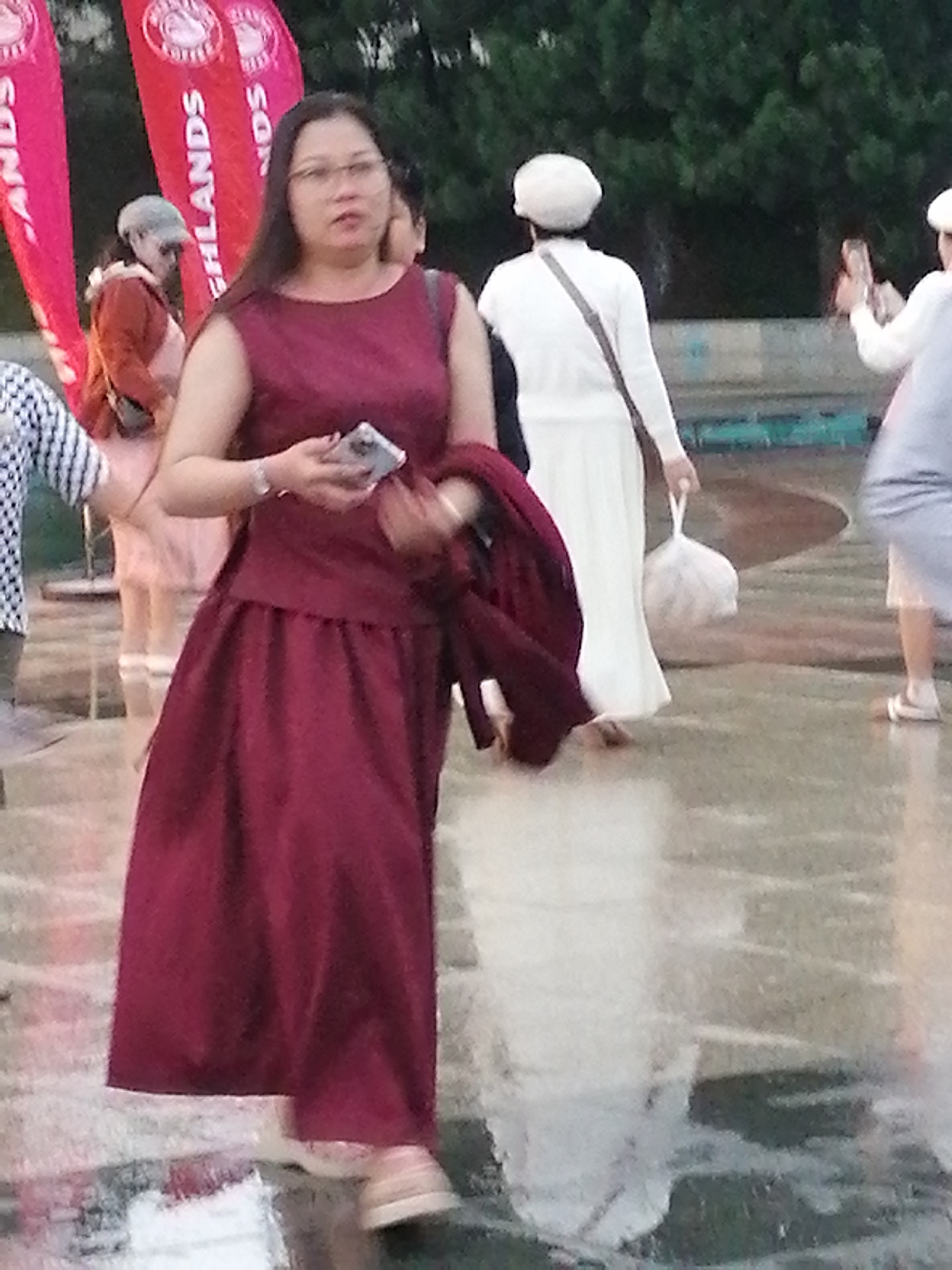 The image shows a group of people outdoors, with a focus on a woman in the foreground wearing a maroon dress and glasses. She is holding a smartphone in her right hand and appears to be looking towards the right of the frame. Behind her, other individuals are visible, some wearing white clothing. In the background, there are trees and large red banners with text, one of which reads "HIGHLANDS COFFEE". The ground appears wet, suggesting it may have recently rained or is raining. The scene is outdoors, possibly at an event or gathering. The presence of "HIGHLANDS COFFEE" banners suggests a commercial or public event. The overall impression is a candid shot of people in an urban or semi-urban outdoor setting. The context of Da Lat, Vietnam, is not directly evident from the visual information alone but would be provided as external information.
