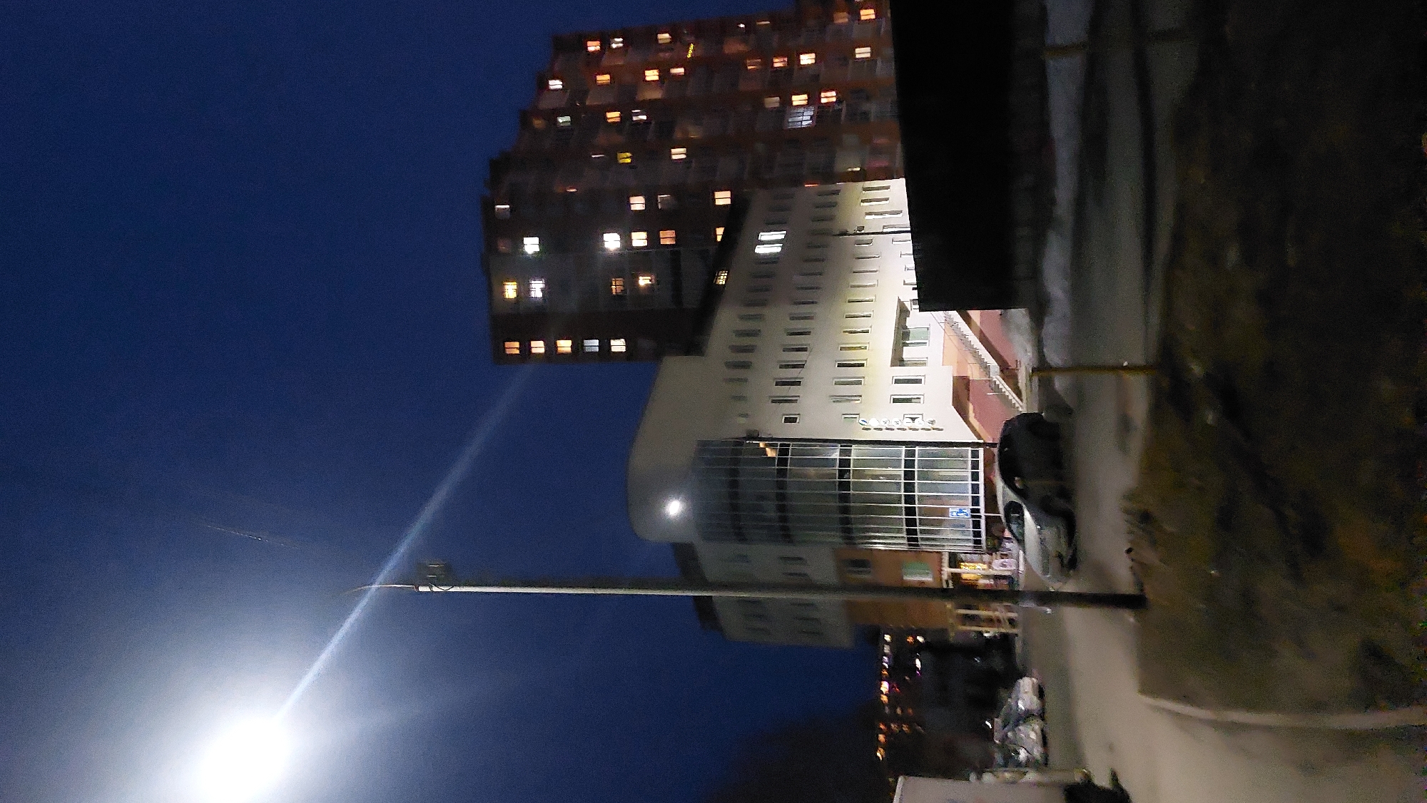The image depicts a night scene in Novosibirsk, Russia. A tall, modern building complex dominates the skyline, with lit windows visible in the higher floors. Below this building stands another building with a glass facade, reflecting the streetlights. In the foreground, a street lamp illuminates the surrounding area, including a sidewalk and parked cars.