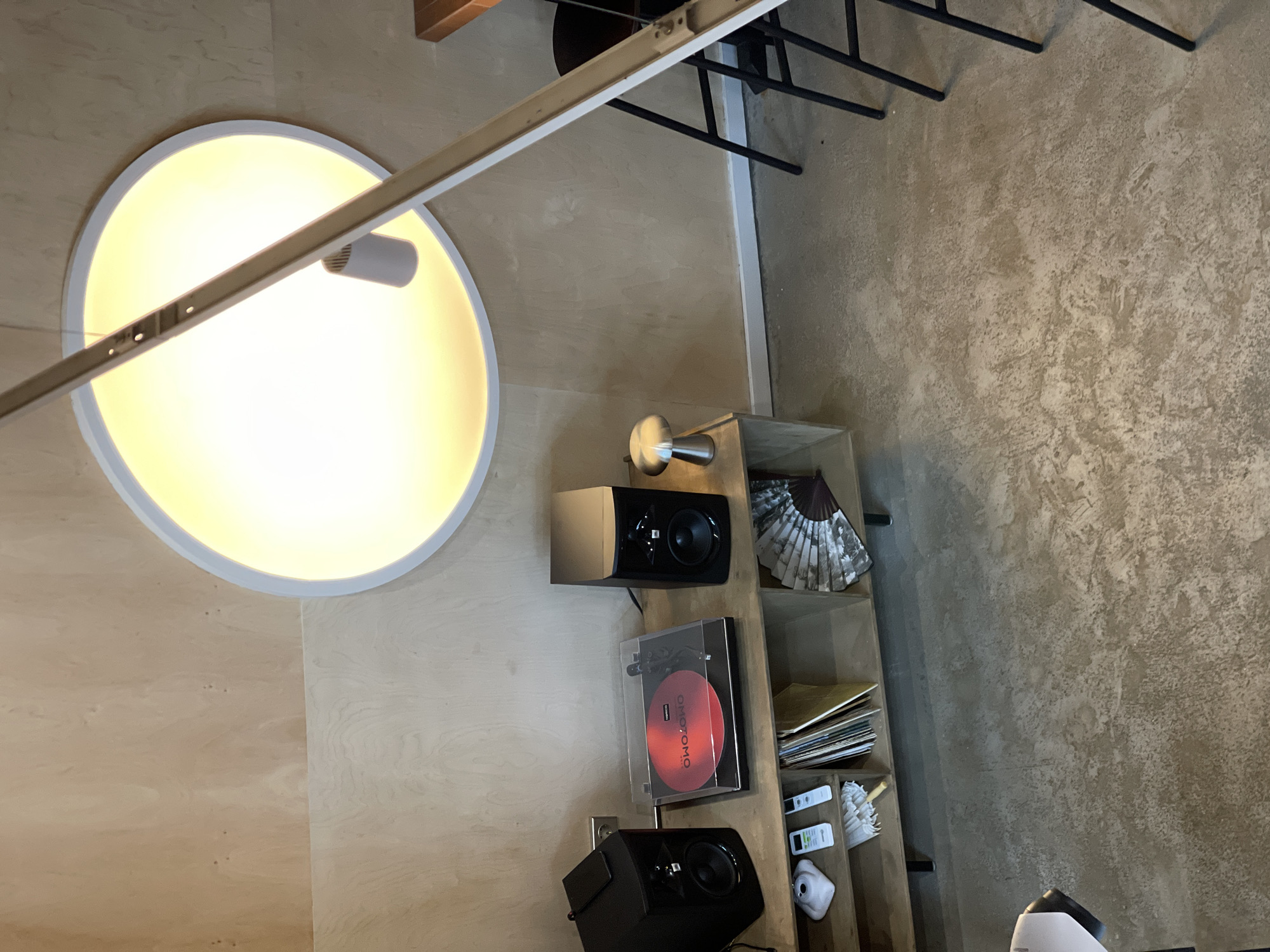 The image depicts an indoor scene, likely a living room or study, with a minimalist aesthetic. The background consists of light-colored wooden walls and a concrete floor. Dominating the upper portion of the image is a large, circular ceiling light with a warm, yellow glow. A long, white fixture hangs from the ceiling, crossing diagonally across the frame and partially obscuring the circular light.
In the mid-ground, a wooden media console is set against the wall. On top of the console, two black speakers flank a record player, which has a red vinyl record spinning on it. To the right of the speakers, a small, silver mushroom-shaped lamp sits on a narrow cabinet. Below the speakers and record player, the console has shelves. One shelf contains several records and a small, white umbrella. Another shelf on the right holds a decorative fan.
In the foreground, the concrete floor is visible, showing variations in texture and color. To the right, the legs of black chairs and a portion of a wooden table are visible, suggesting a dining or seating area nearby.
There are no people present in the image. The scene is static and well-lit, highlighting the arrangement of electronic equipment and decorative items. The overall impression is one of a modern, organized space. There is no information within the image to indicate its location in Izhevsk, Russia.