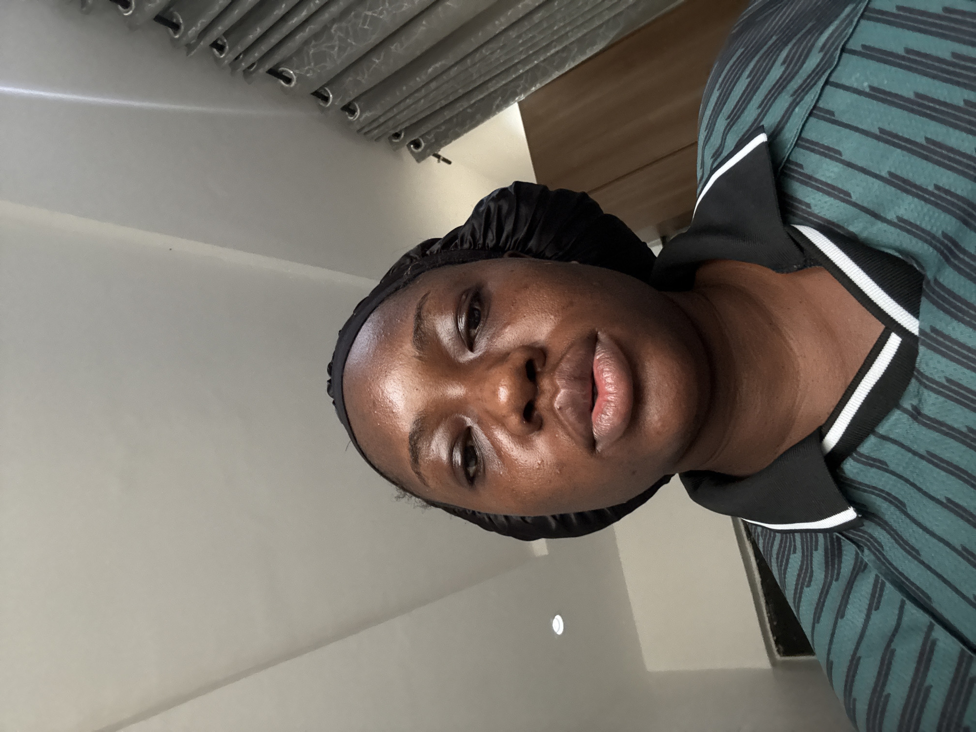 A person wearing a black satin head covering and a green and black striped collared shirt is facing the camera. The background includes a gray curtain, a white wall, and a wooden door. There is no discernible indication of Iddo Tudunwada, Nigeria.