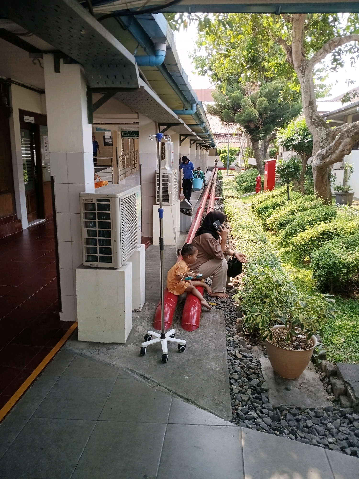 The image depicts an outdoor hospital corridor in Purwodadi, Indonesia. In the foreground, a child wearing an orange shirt sits on a bright red medical apparatus, possibly a leg support, while a woman wearing a hijab and brown attire sits beside them, engaged in a phone conversation. To the left, a white air conditioning unit is mounted on a pedestal. Further down the corridor, an IV pole with a drip bag stands near another air conditioning unit. Beyond the corridor, a lush green garden with various trees and plants is visible, with a red fire hydrant and a potted plant. A person in a blue shirt carrying a blue bucket walks away in the distance. The architecture suggests a tropical climate with an open-air walkway.