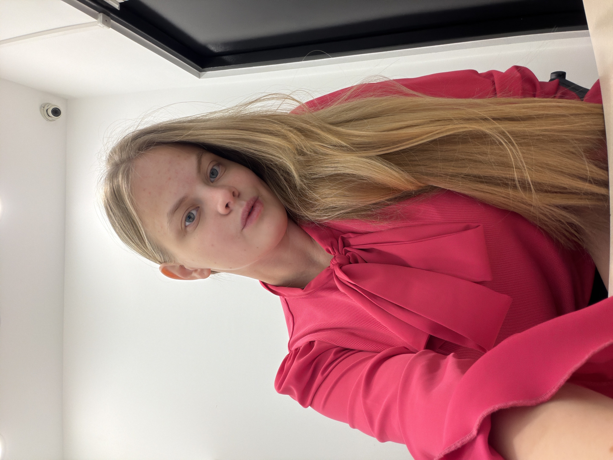 A woman with long blonde hair is facing the camera. She is wearing a bright pink long-sleeved blouse with a large bow at the neck. Behind her is a white wall with a security camera in the upper left corner. Above the woman is a black rectangular object, possibly a light fixture. The location is not discernable as Izhevsk, Russia.