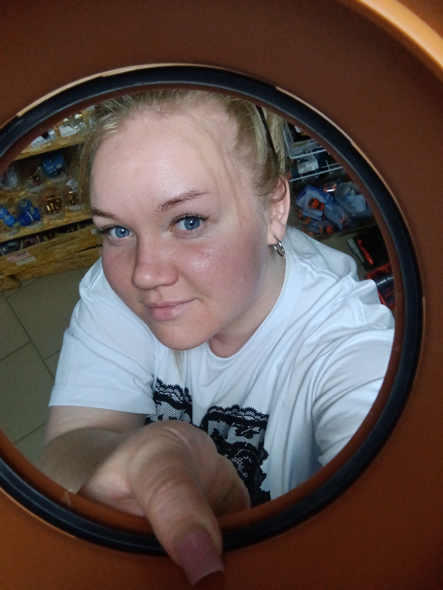 A woman with blonde hair and blue eyes is looking up into the camera through a large circular orange-colored plastic opening. She is wearing a white t-shirt with a black lace pattern on it. Her left hand is extended towards the camera. Shelves with various hardware supplies and tools are visible in the background. The image appears to be taken indoors in a retail or storage area. There is no information in the image to determine if the location is in the city of Фоки, Russia.