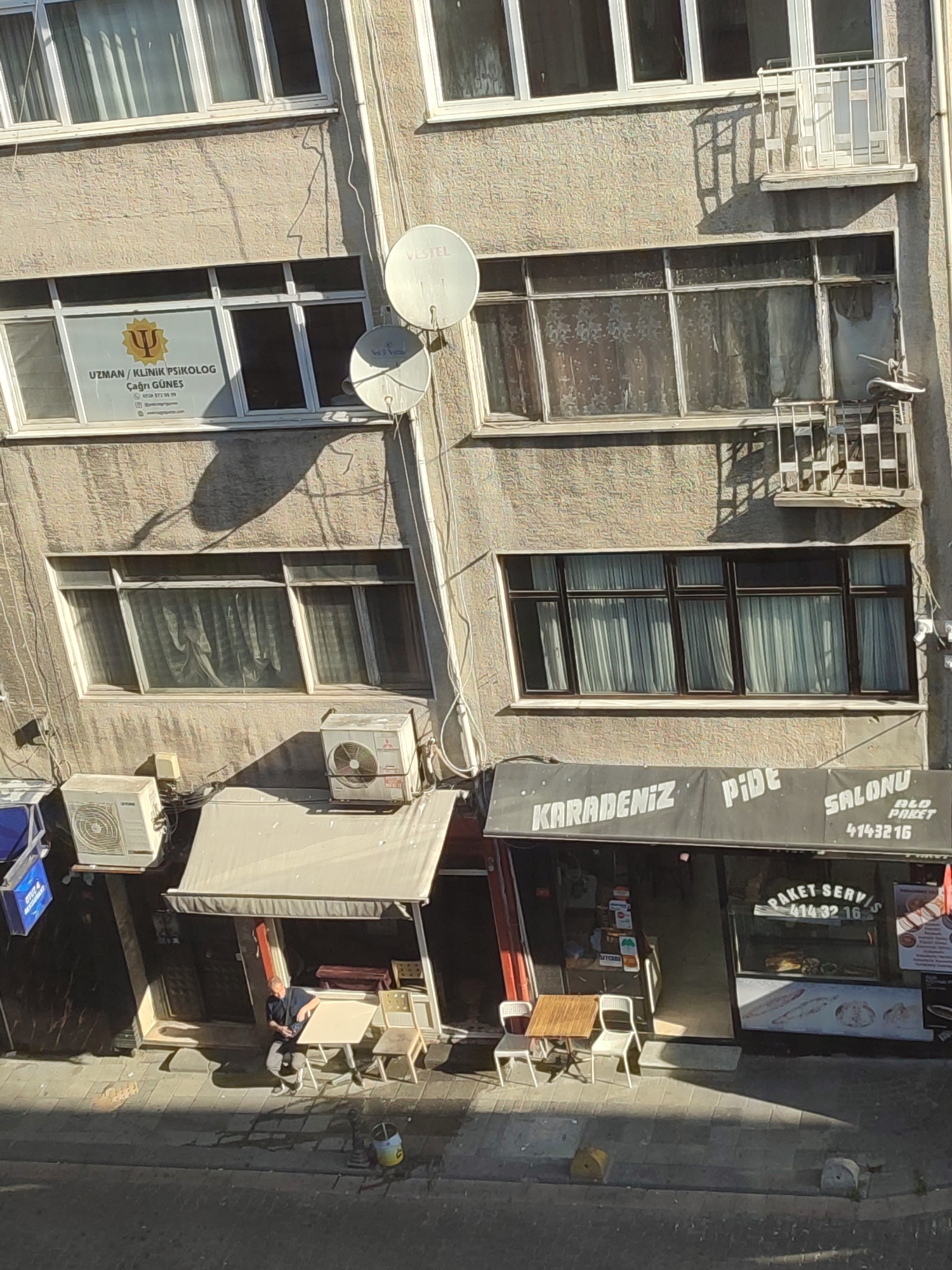 The image shows a street view of a building in Kadıköy, Turkey. On the first floor, there are two businesses. The one on the left is a restaurant with an awning and tables and chairs outside. A man is sitting at one of the tables. The business on the right is also a restaurant, with a sign that says "KARADENIZ PIDE SALONU". Inside, food is visible in a display case. Above the businesses, there are several windows and air conditioning units on the building's facade. Two satellite dishes are mounted on the wall.