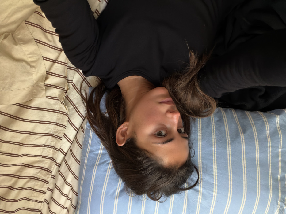 A female individual is depicted lying on her left side on a bed. Her head rests on a light blue pillow with thin white horizontal stripes. Her long, dark brown hair is spread across the pillow and partially covers her right shoulder. She is wearing a long-sleeved black top. Her face is directed slightly towards the viewer, with her eyes open. The skin on her face exhibits a natural, warm tone. The bed is covered with bedding featuring a beige or cream color with thin brown vertical stripes. The upper part of her right arm is visible, bent at the elbow, and extends towards the upper right of the frame, suggesting she may be holding a device for self-photography. The scene is indoors, consistent with a private residential setting. The specific location within Paris, France, cannot be determined from the visual information provided in the image.