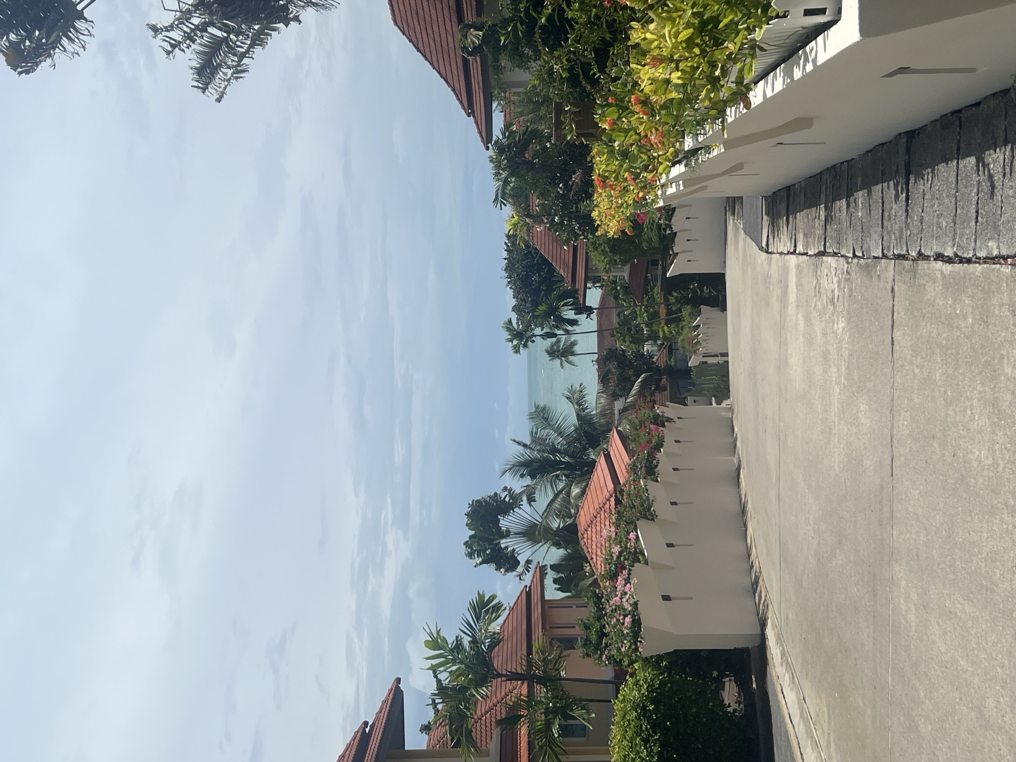 The image depicts a paved road winding downhill towards a tropical coastline, likely within Ko Samui District, Thailand. On either side of the road are white walls topped with decorative crenellations, behind which are visible houses with red tiled roofs and lush green foliage, including palm trees. The sea is visible in the distance, under a hazy, overcast sky. There are no people or animals present in the scene.