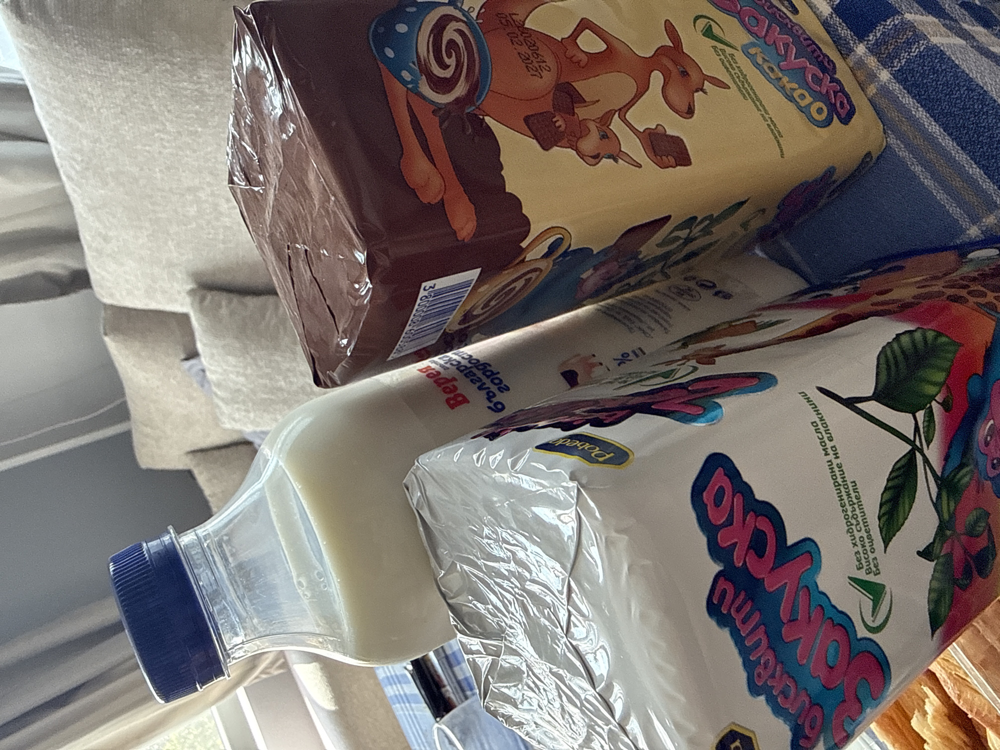 The image displays three food products – two packaged snacks/biscuits and one bottle of milk – arranged on a blue and white striped fabric. The background features light-colored upholstered furniture, indicating an indoor setting in Rogosh, Bulgaria. The bright, natural lighting suggests it is daytime.
The top product package is yellowish-tan, featuring a cartoon illustration of a kangaroo-like animal with a blue-spotted pattern on its back, carrying a smaller creature. Visible text from OCR includes "Закуска" (Breakfast), "какао" (cocoa), "Без хидрогенирани масла" (No hydrogenated oils), "Високо съдържание на влакнини" (High fiber content), and "Без оцветители" (No artificial colors). A batch number "L28020612" and a partial date "05.02.202" are present, along with a barcode starting with "3800008".
Adjacent to this, a white packaged product, partially covered in crinkled clear plastic, displays colorful text and an illustration of a plant with green leaves and reddish elements. This package, identified as "Pobeda" brand "Закуска бисквити" (Breakfast biscuits), also notes "Без оцветители" (No artificial colors), "Без хидрогенирани масла" (No hydrogenated oils), and "Високо съдържание на влакнини" (High fiber content).
A clear plastic bottle with a blue screw cap contains white milk. Its label prominently shows the brand "Верея" and the phrase "българска гордост" (Bulgarian pride). The OCR also detected "2.0% захар" (2.0% sugar) on the bottle. All items appear new and unopened.