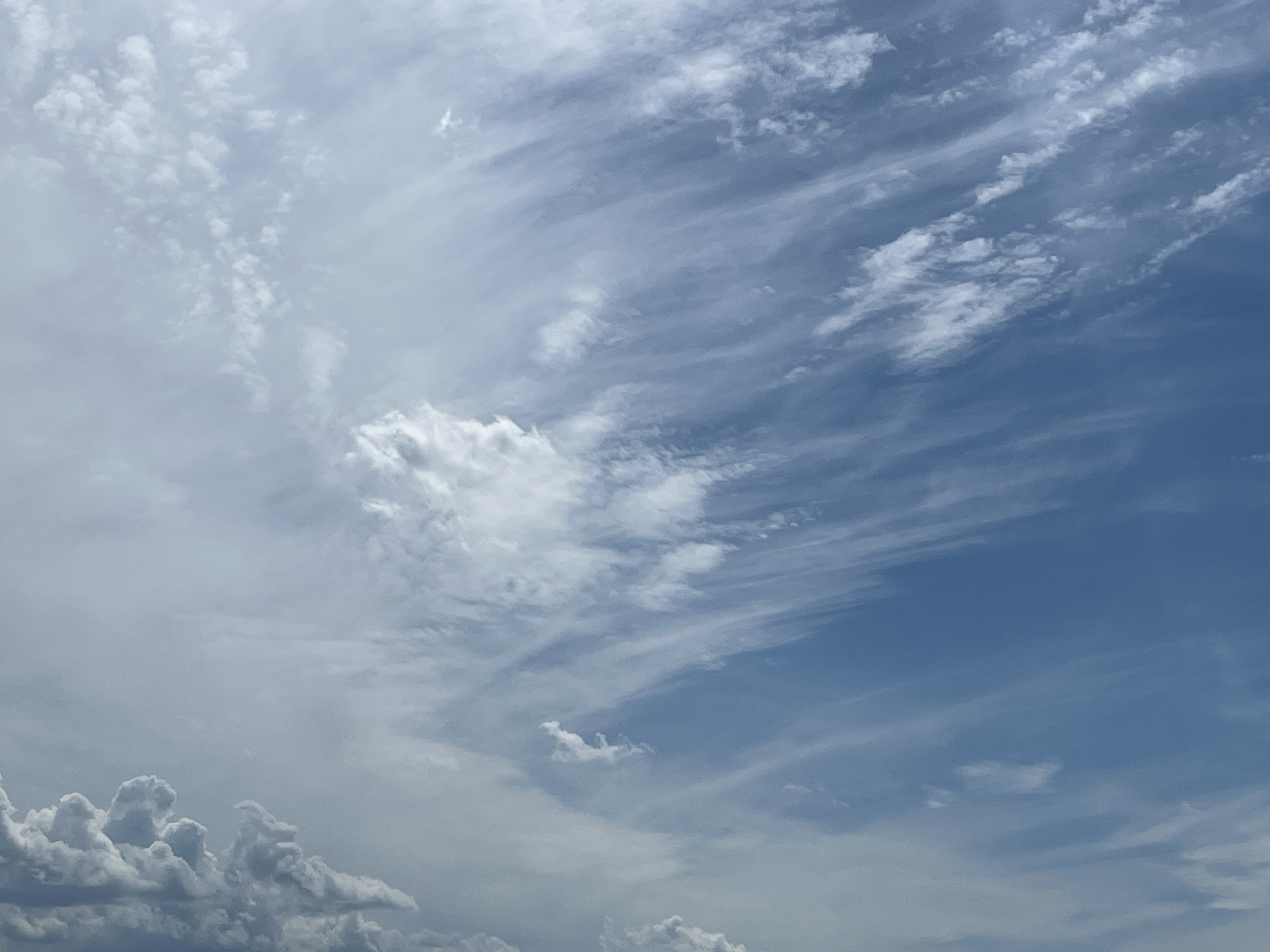 The image depicts a sky filled with a mixture of cloud types. Wispy cirrus clouds stretch across the upper right portion of the frame, blending into more textured cumulus clouds that occupy the left and lower sections. The sky transitions from a bright, hazy blue on the right to a paler, overcast grey on the left, suggesting varied atmospheric conditions. There are no people, structures, or ground visible in the image. The scene is devoid of any specific landmarks that would indicate its location within Yekaterinburg, Russia.