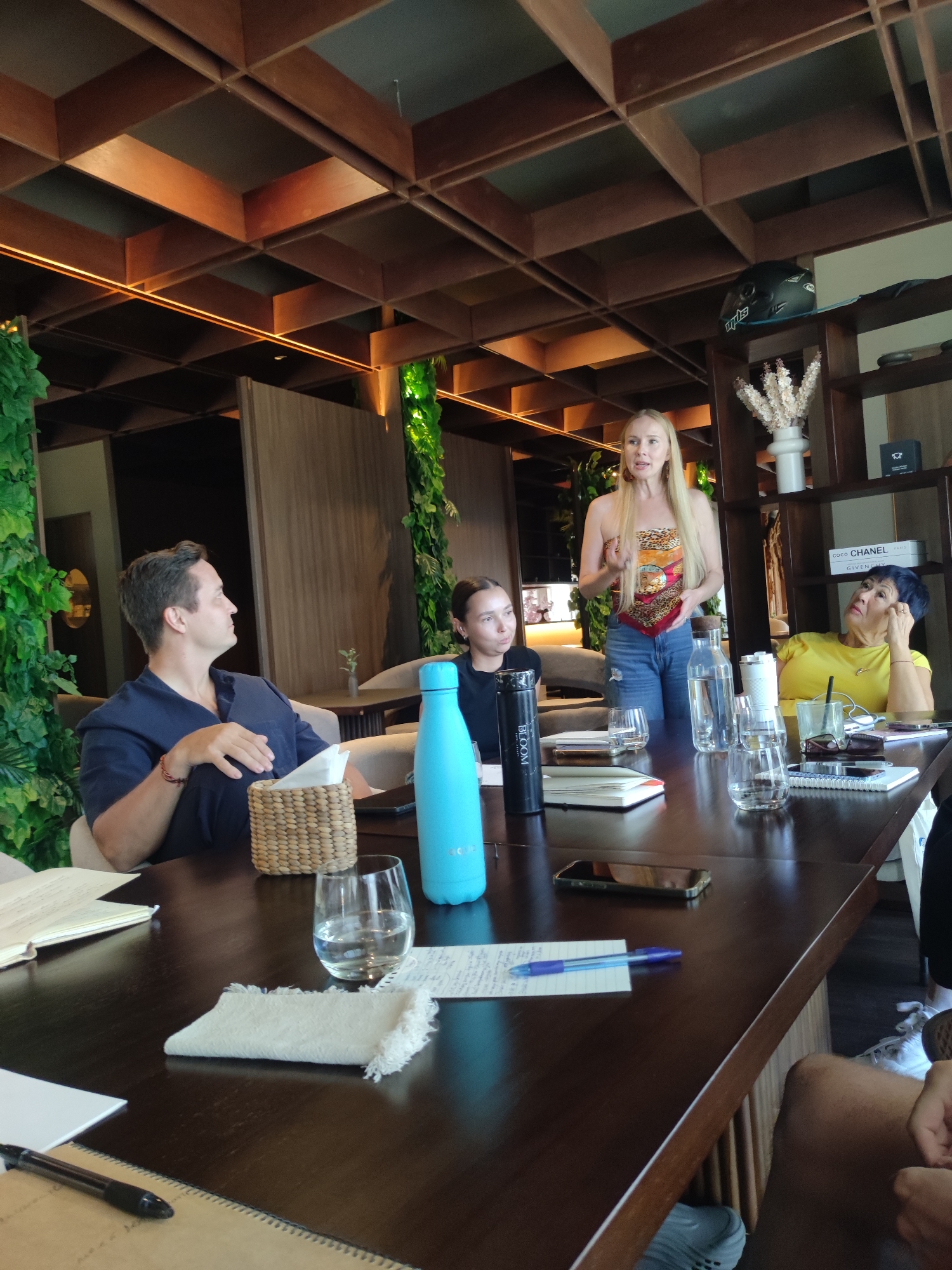 A group of four individuals is gathered around a dark brown wooden table in what appears to be a restaurant or a meeting space in North Kuta, Indonesia. The space is characterized by a prominent wooden ceiling lattice and vertical wall panels decorated with green foliage.
One woman with long blonde hair, wearing a patterned top and jeans, stands at the table and is speaking or presenting to the others. To her left, a younger woman in a black shirt listens attentively, with a black water bottle in front of her. To the right of the standing woman, another woman in a yellow shirt leans with her head propped on her hand, listening. A man in a dark blue shirt sits to the left of the younger woman, also paying attention.
The table has various items on it, including water bottles, glasses, notebooks, pens, cell phones, and a small woven basket. A white cloth napkin is visible near a water glass. There is written material on a piece of paper under a pen.
In the background, a shelf unit holds decorative items, including a black motorcycle helmet, white floral arrangements, and some boxes with names on them. The overall atmosphere appears to be professional and attentive.