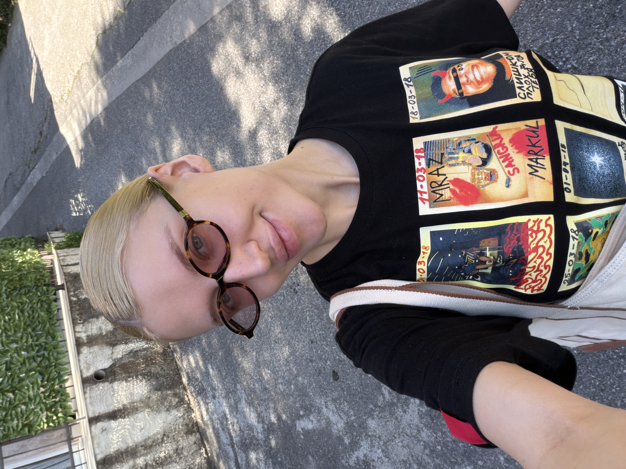 A high-angle outdoor selfie captures a person with short, slicked-back blonde hair, wearing round, tortoise-shell framed glasses with tinted lenses. The person looks directly at the camera with a neutral, slightly smirking expression. They are dressed in a black short-sleeved t-shirt featuring multiple square graphics resembling album covers or concert posters. A cream and brown striped shoulder bag strap is visible across their chest.
Notable text on the t-shirt graphics includes dates such as "18-03-18", "11-03-18", "01-04-18", and "25-03-18". One graphic depicts a man with devil horns and sunglasses, accompanied by Cyrillic text reading "СЛИШКОМ ПЛОХ ОЛЯ ТЕБЯ" (translated as "Too bad, Olya, for you" or "Too bad for you, Olya"). Other discernible words on the graphics include "MRZ", "SANGELIY", and "MARKUL".
The background features grey asphalt or concrete ground, brightly lit by sunlight which casts prominent shadows, indicating a clear, sunny daytime. To the left, a wall topped with dense green foliage is visible. The scene suggests an outdoor urban or suburban setting, specifically Medinski Krš, Montenegro, based on the provided context. The person appears to be taking a self-portrait.