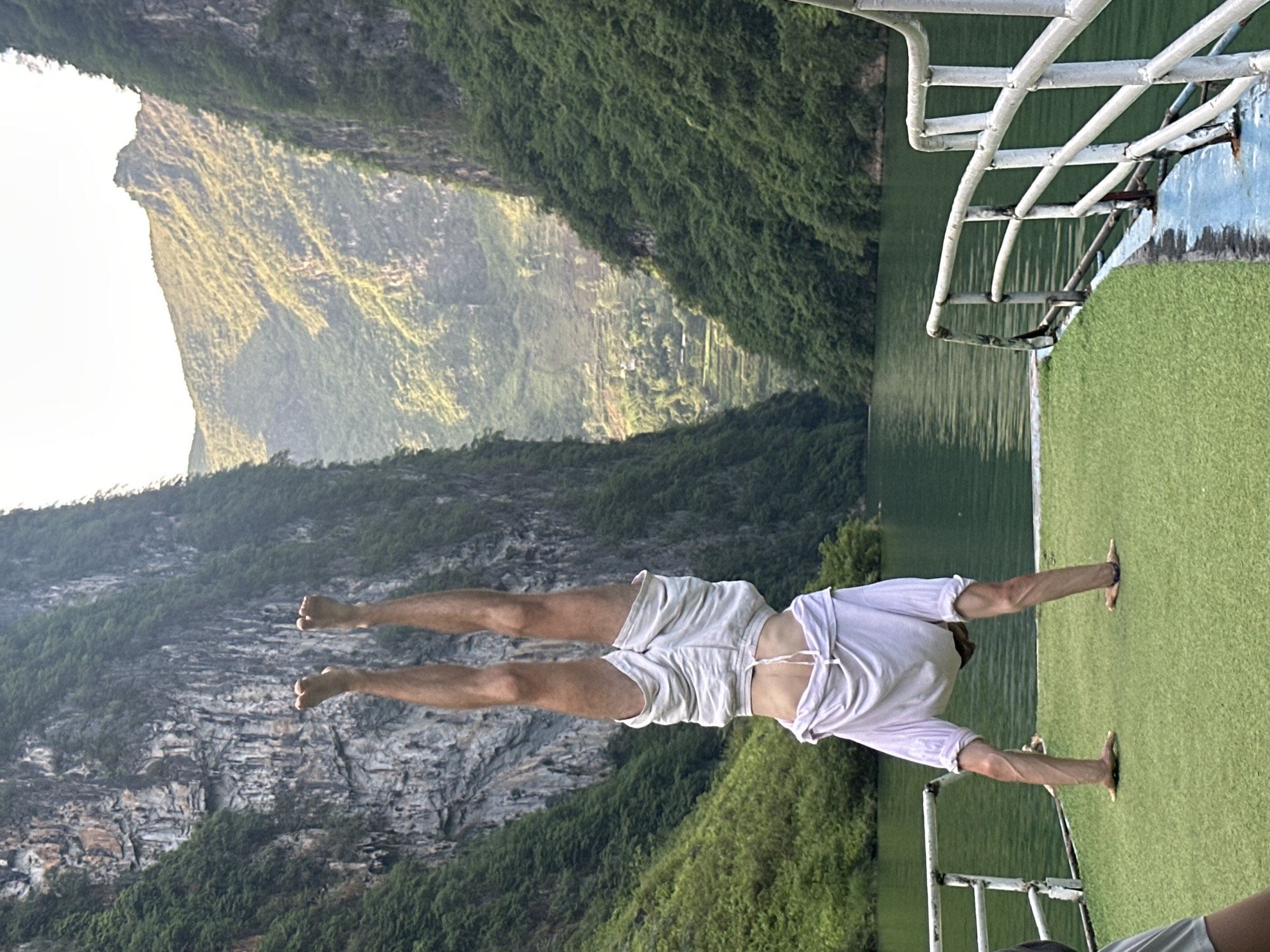 A man in a white t-shirt and light-colored shorts is performing a handstand on a boat, with his feet facing towards a mountainous landscape. The boat has a green artificial turf surface and white metal railings. The mountains are covered in lush greenery, and a body of water is visible next to the boat. The scene appears to be located in a mountainous area, possibly near the Đồng Văn Commune in Vietnam.