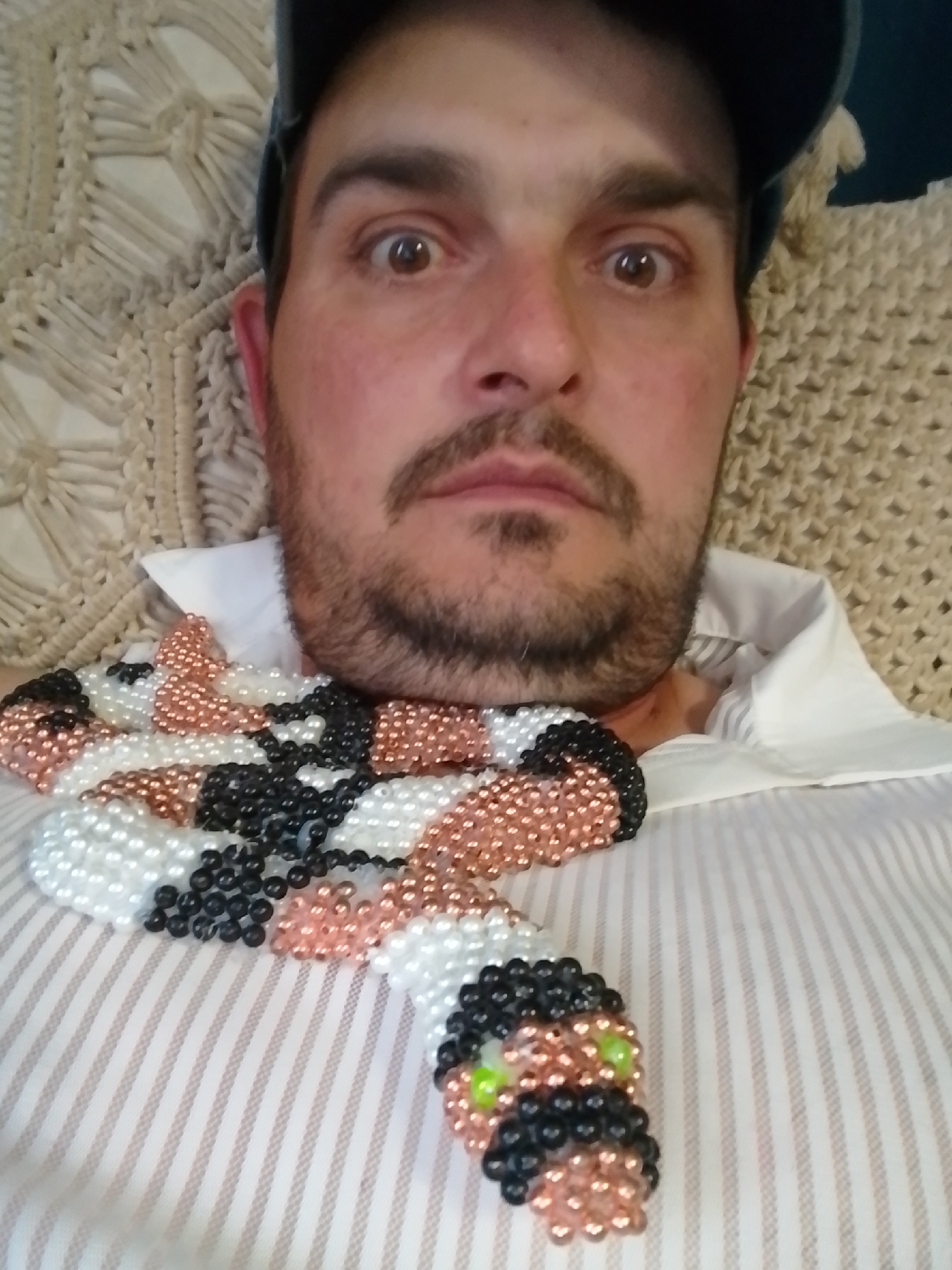 The image shows a close-up, overhead, selfie-style shot of a man wearing a white, pinstriped shirt and a dark baseball cap. A beaded snake accessory, made of black, white, and copper beads, is draped around his neck and rests on his shirt. The snake's eyes are depicted with green beads. The man has brown eyes, dark hair, and a beard and mustache. He appears to be lying down on a textured, woven surface, possibly a blanket or cushion, with a macrame-like pattern. The background is soft and out of focus. The location is not specified in the image or provided context.